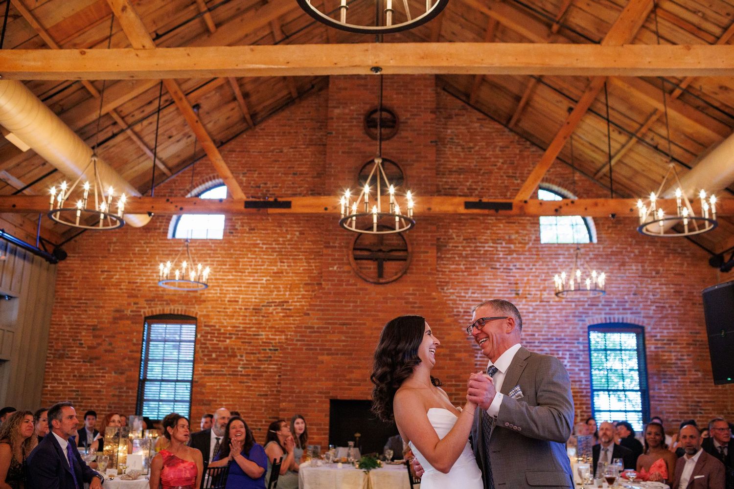 Bride shares emotional father-daughter dance in elegant brick venue with chandeliers and guests watching.