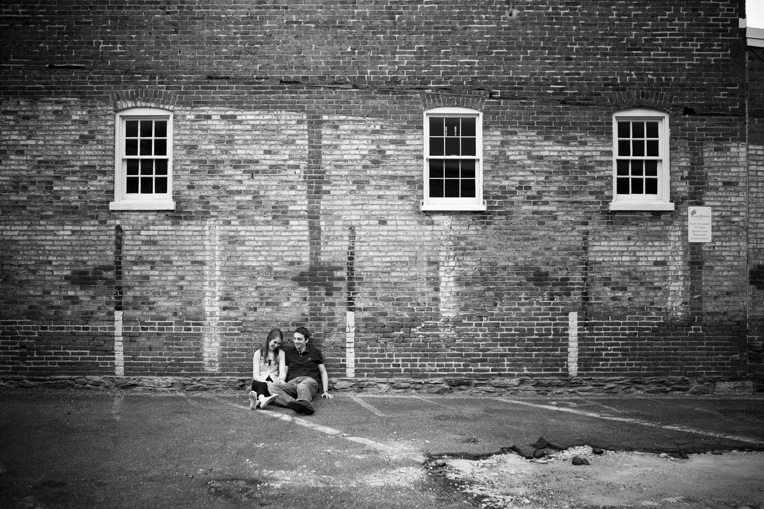 A black and white photo of a brick wall with three windows and people sitting below.