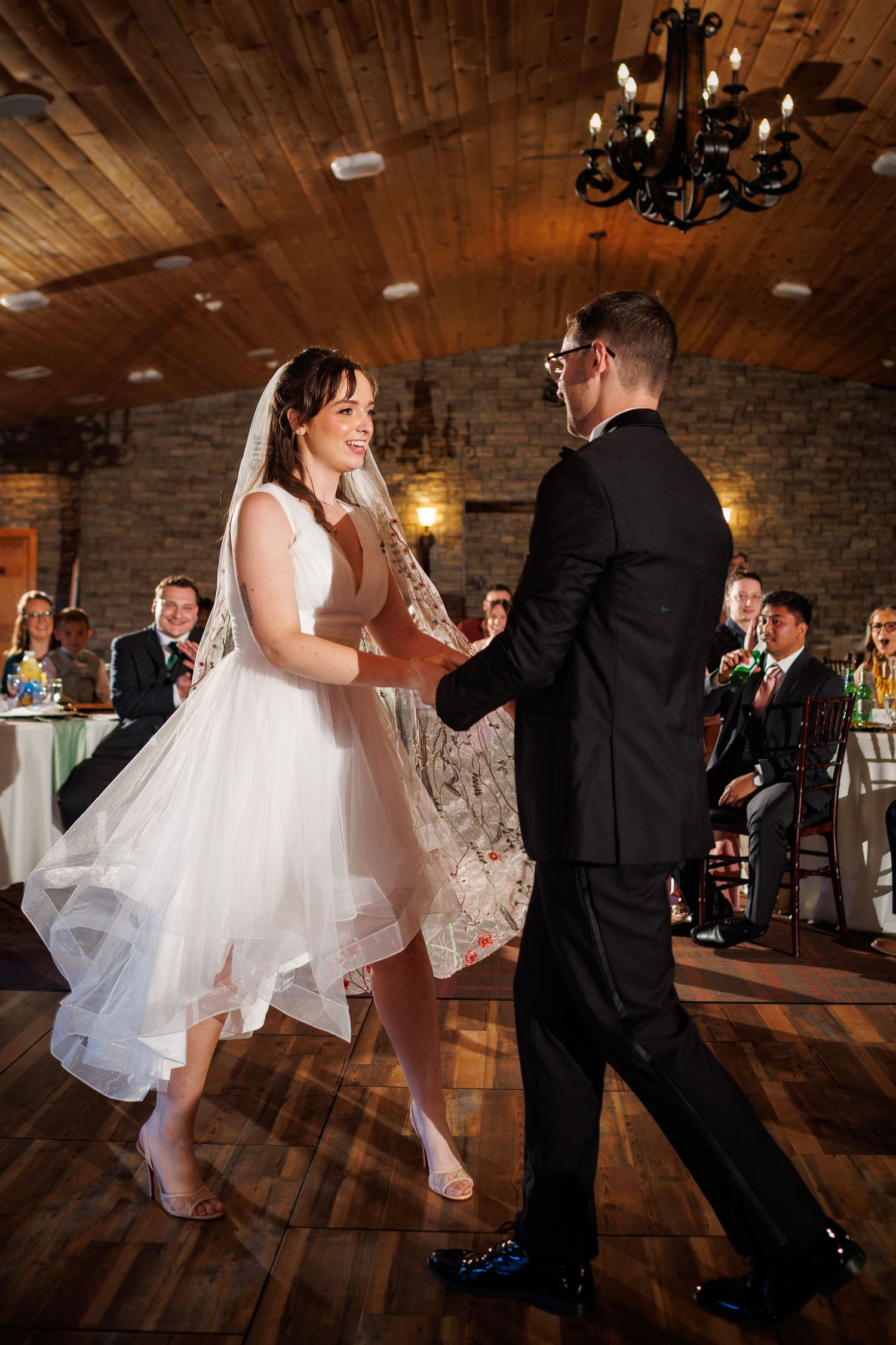 Bride and groom share first dance in rustic wood-paneled reception hall with guests watching.