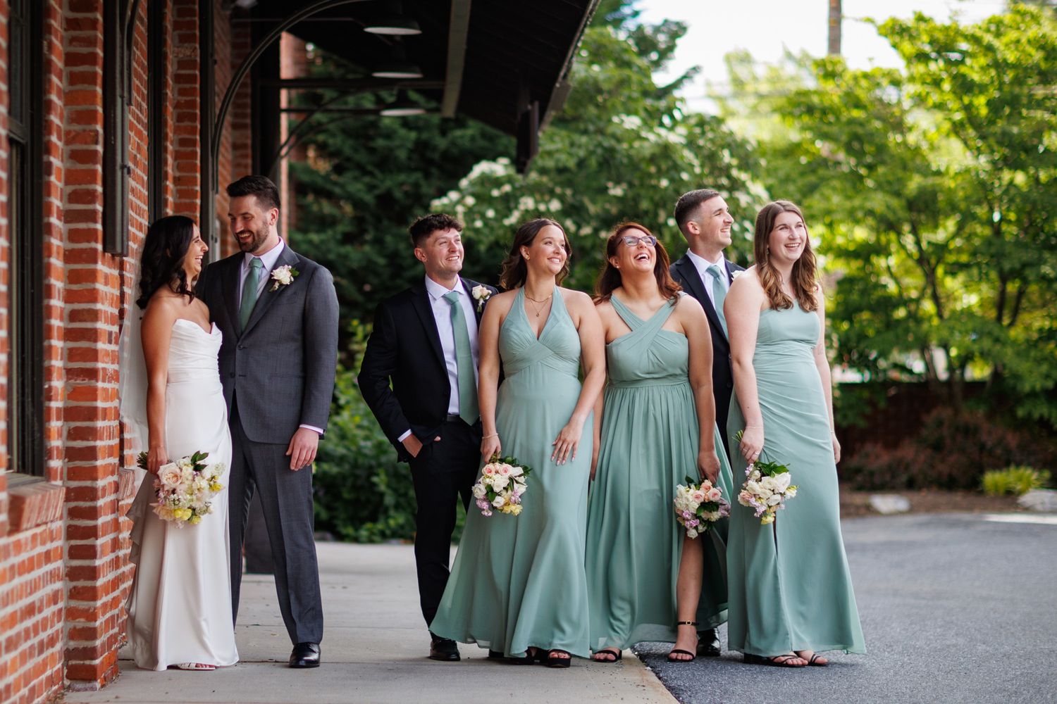 Wedding party in sage green bridesmaid dresses and suits walking outside a brick building with lush greenery.