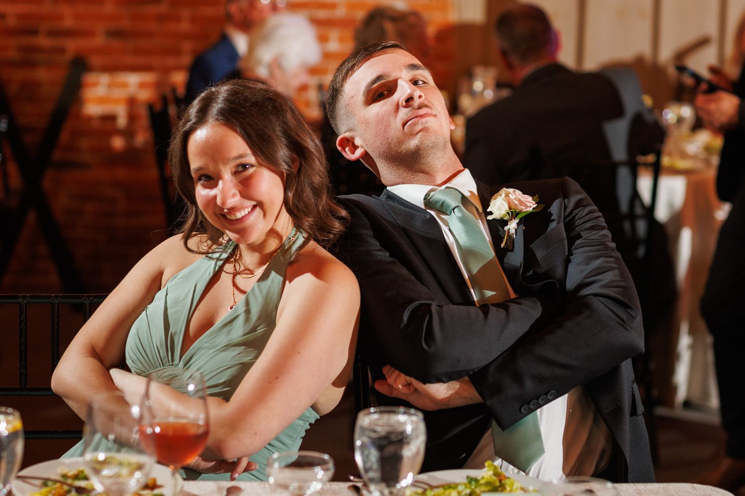 Bride and groom sitting back-to-back at wedding reception table, laughing in a rustic brick venue.