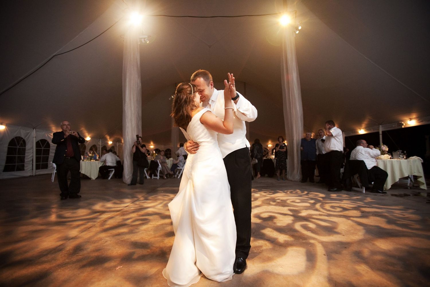 A couple shares their first dance under glowing lights in a reception tent with patterned floor projections.
