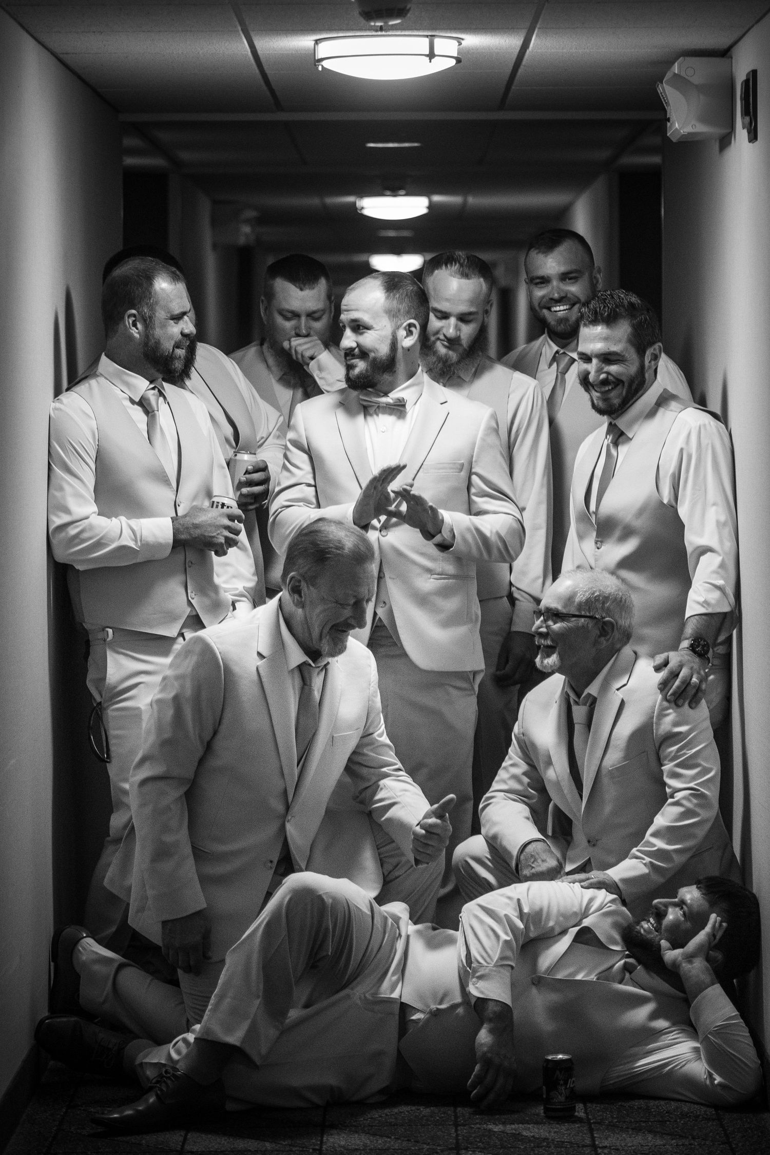 A group of groomsmen in light-colored suits gather around looking at something on a phone in a hallway.