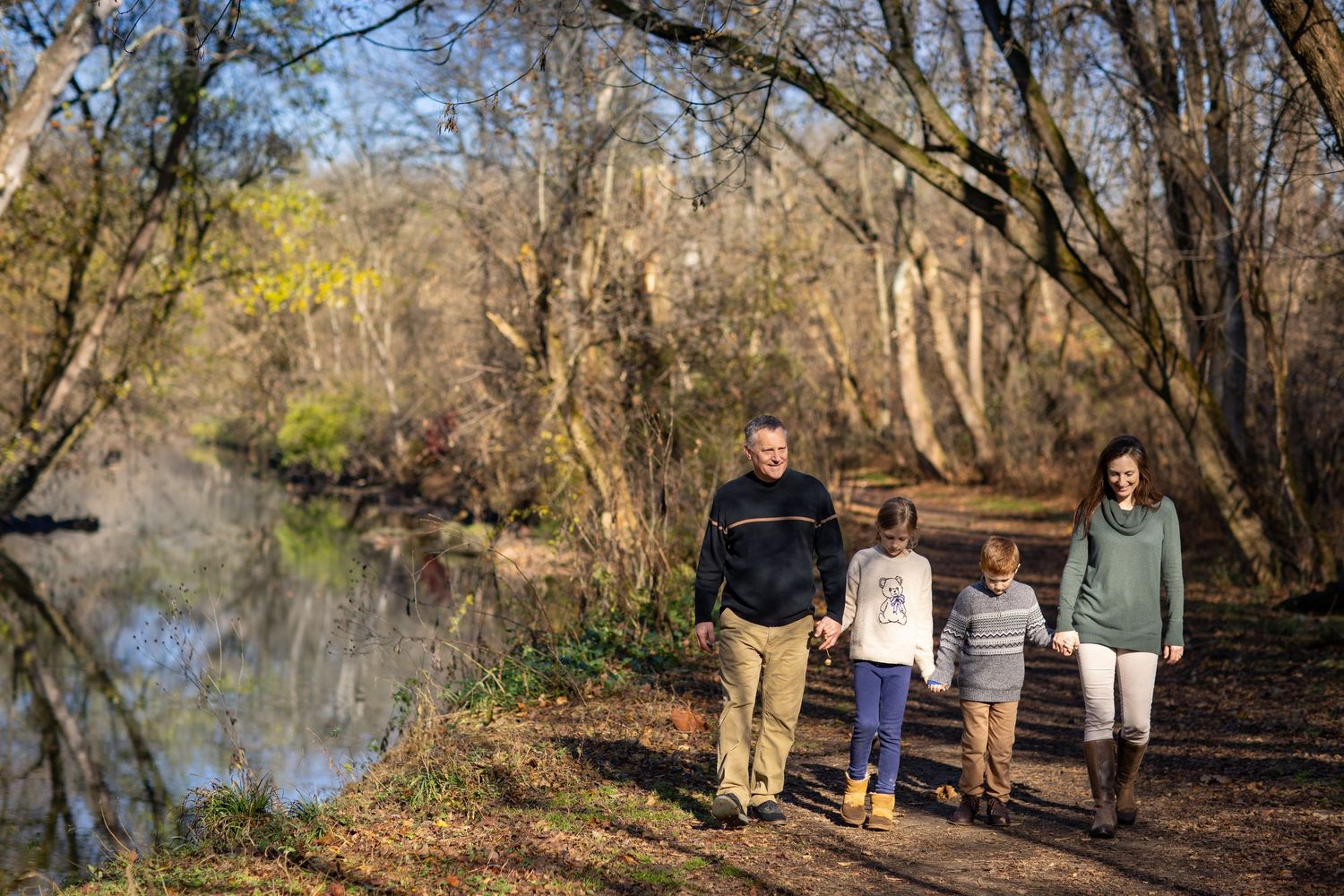 A group walks together on a scenic trail alongside a peaceful river on an autumn afternoon.
