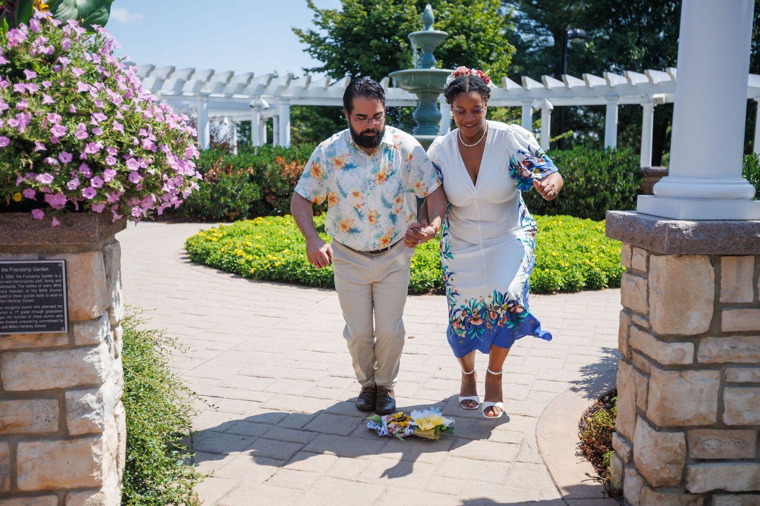 Couple walking hand-in-hand through a beautiful garden pergola surrounded by colorful flowers on a sunny day.