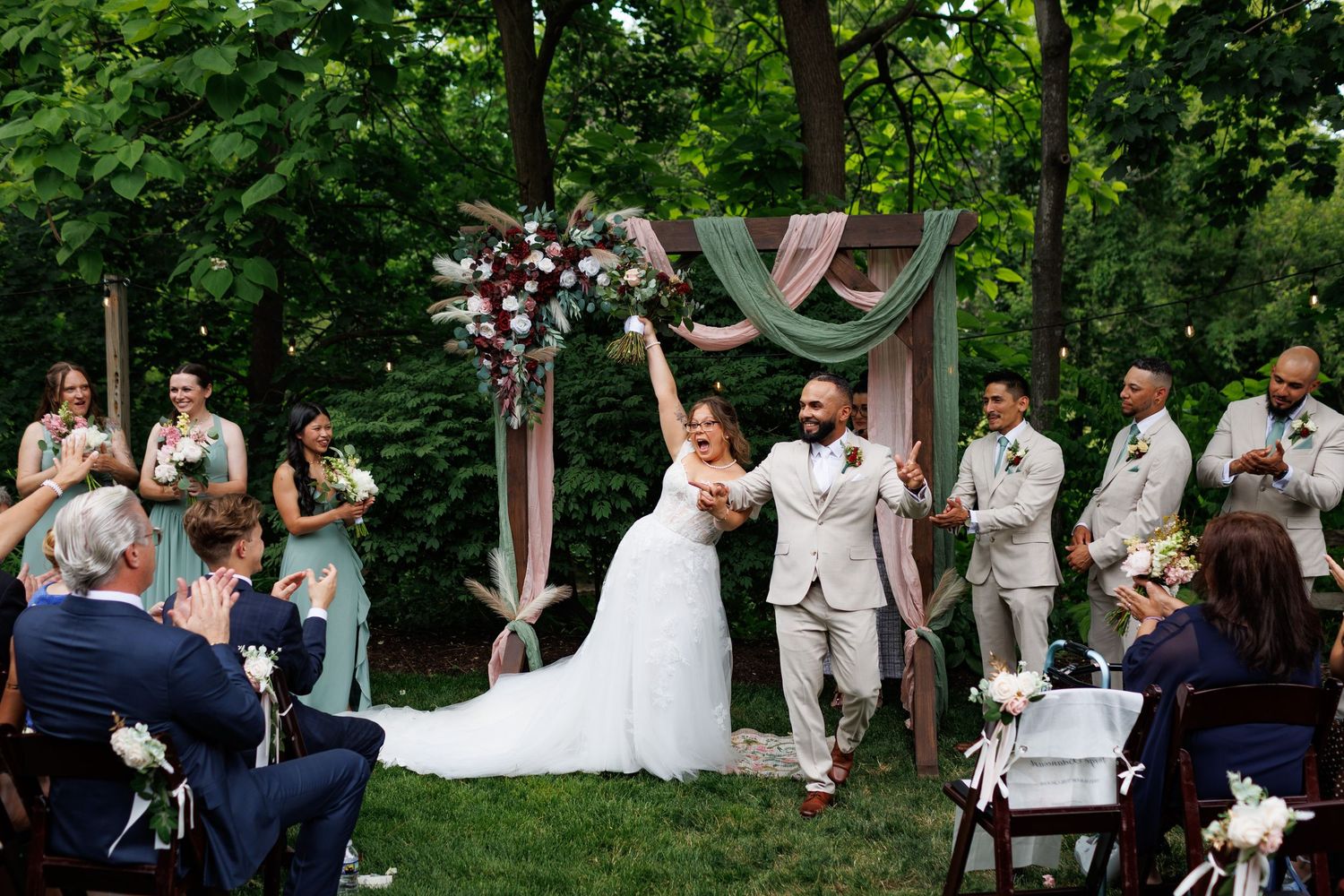 Joyful newlyweds celebrate at outdoor wedding ceremony with bridal party under floral arch surrounded by lush greenery.