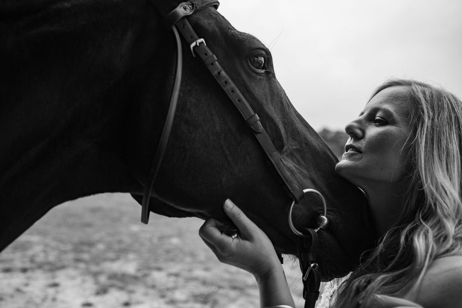 Black and white photo of a blonde woman tenderly kissing a dark horse on the nose outdoors.