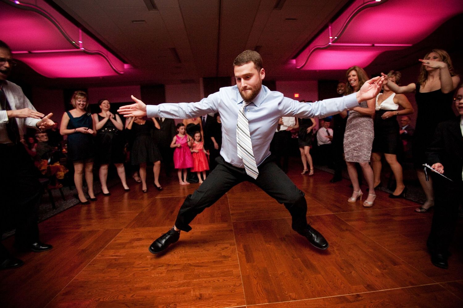 People dance on a wooden floor under pink lighting during a dance party or celebration.
