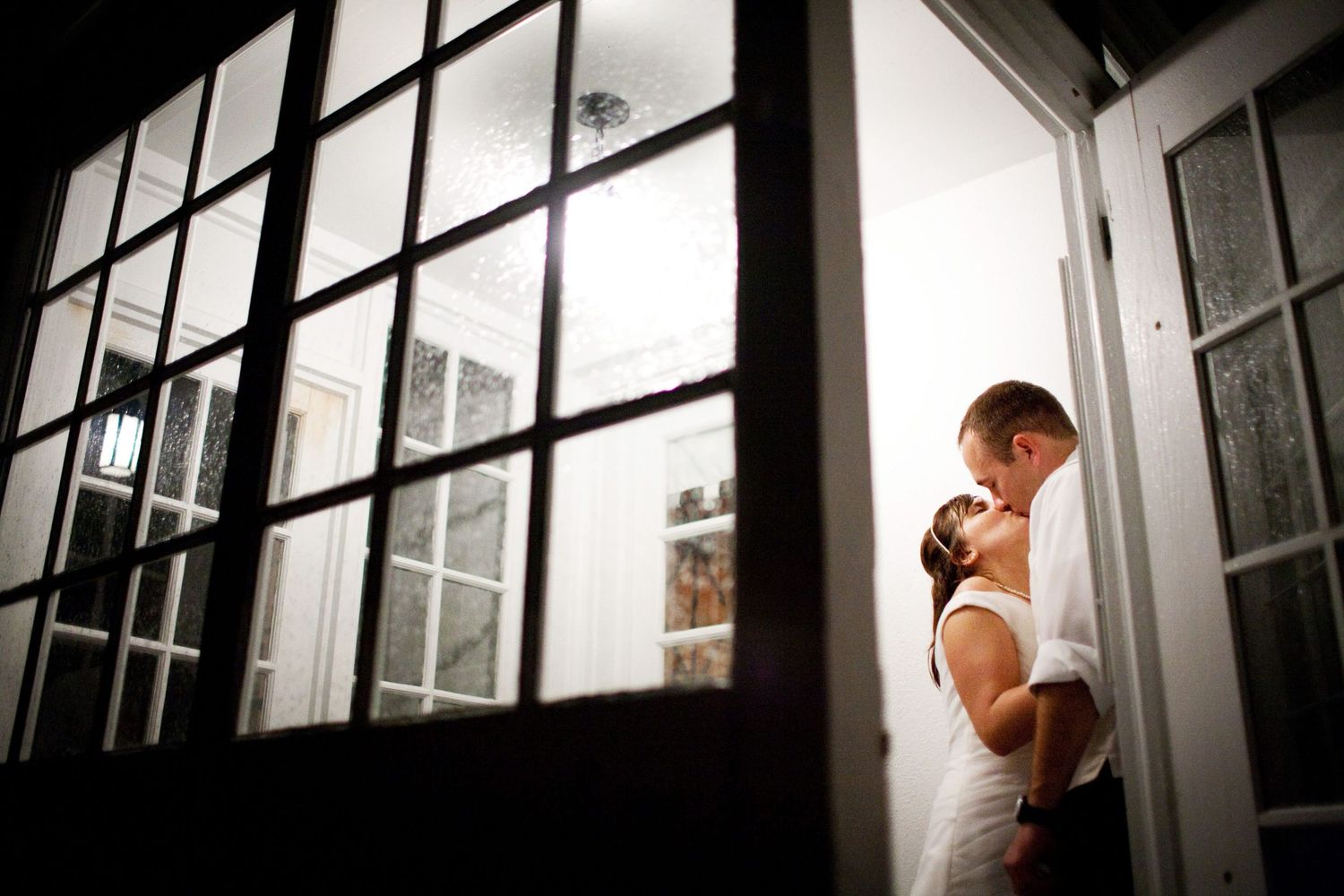Two people share an intimate moment in the doorway of a house with dramatic window lighting casting shadows.