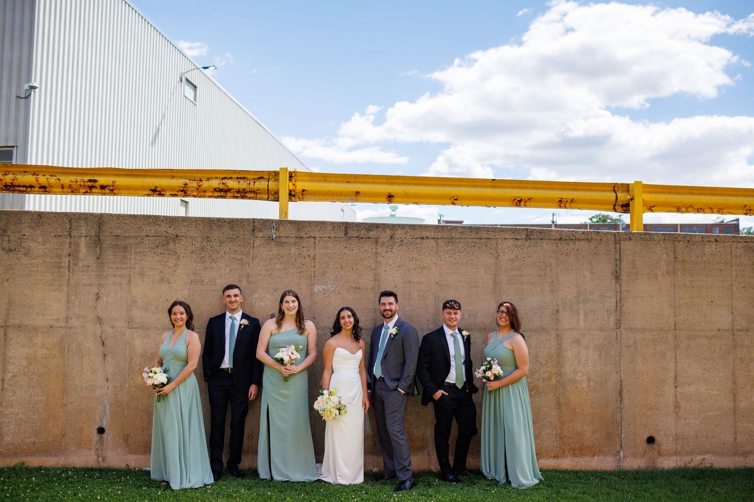 Wedding party poses outdoors in sage green bridesmaid dresses and suits against a rustic concrete wall.