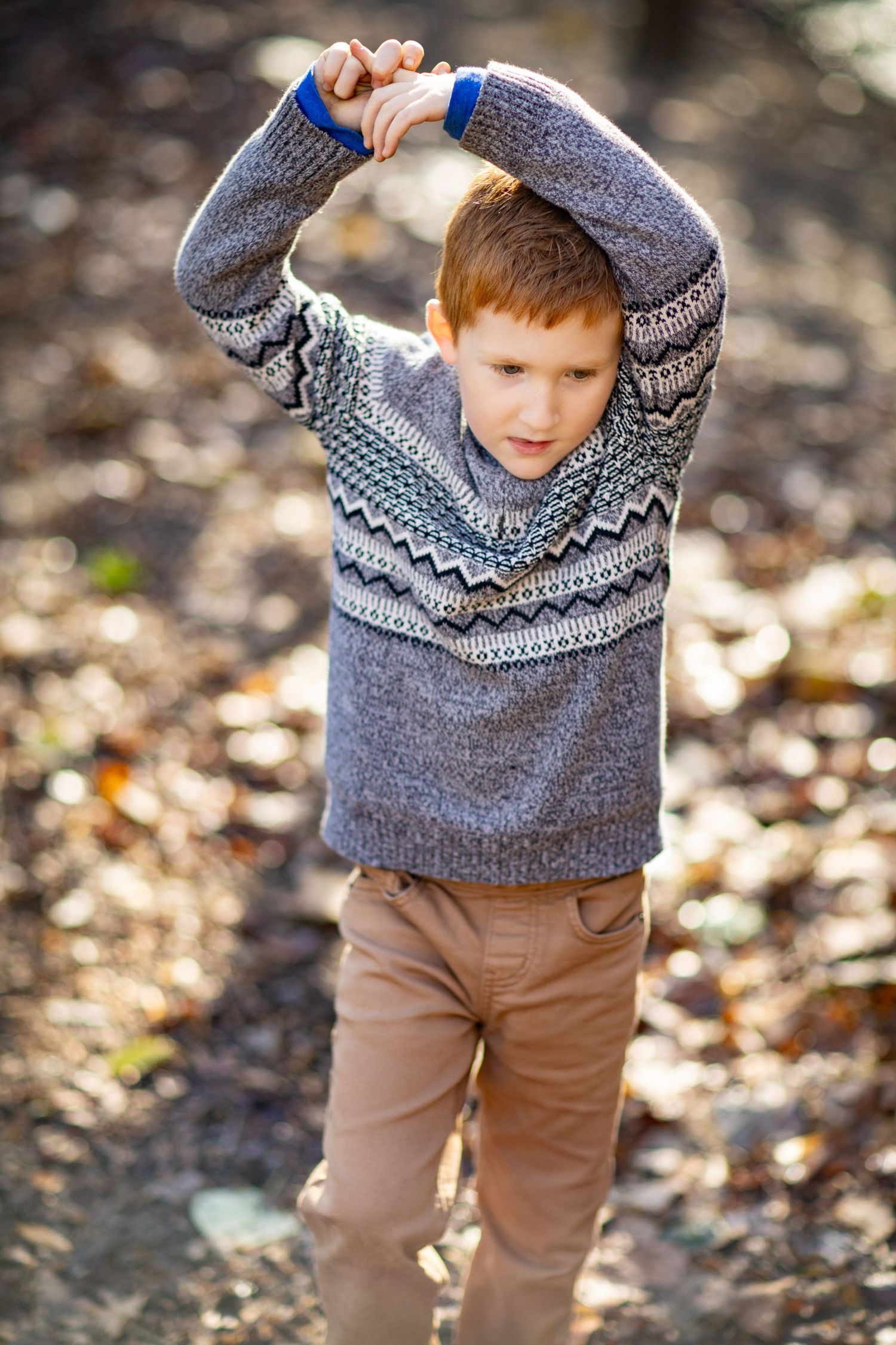 Child in gray patterned sweater and brown pants plays outdoors in autumn setting.