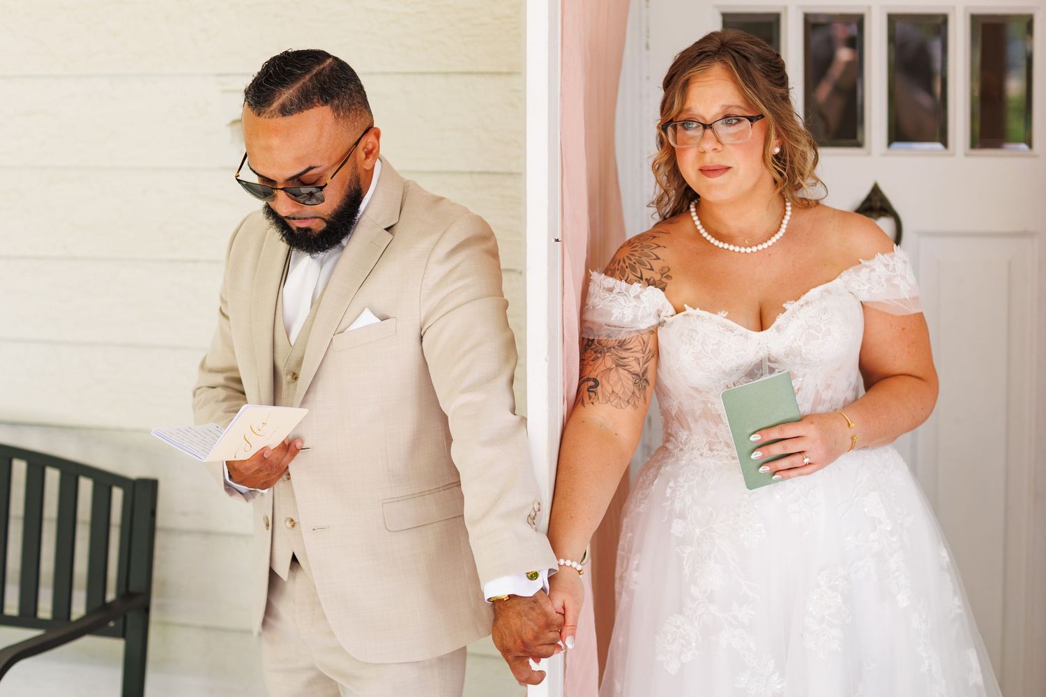 Bride and groom share a tender first look moment, holding hands while reading vows outside a white building.