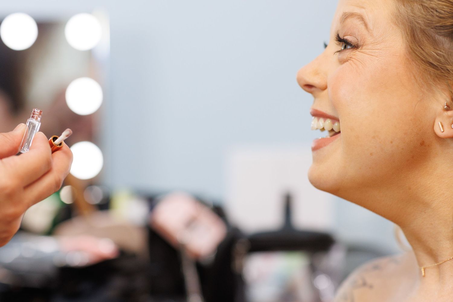 Bride smiling as makeup artist applies lipstick in a brightly lit vanity mirror setting.
