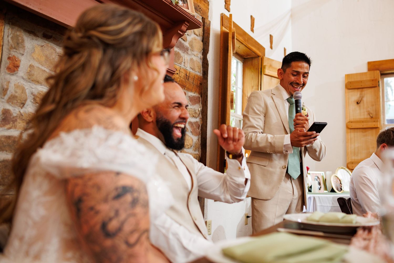 Tattooed groom laughing with best man giving speech at rustic stone venue wedding reception.