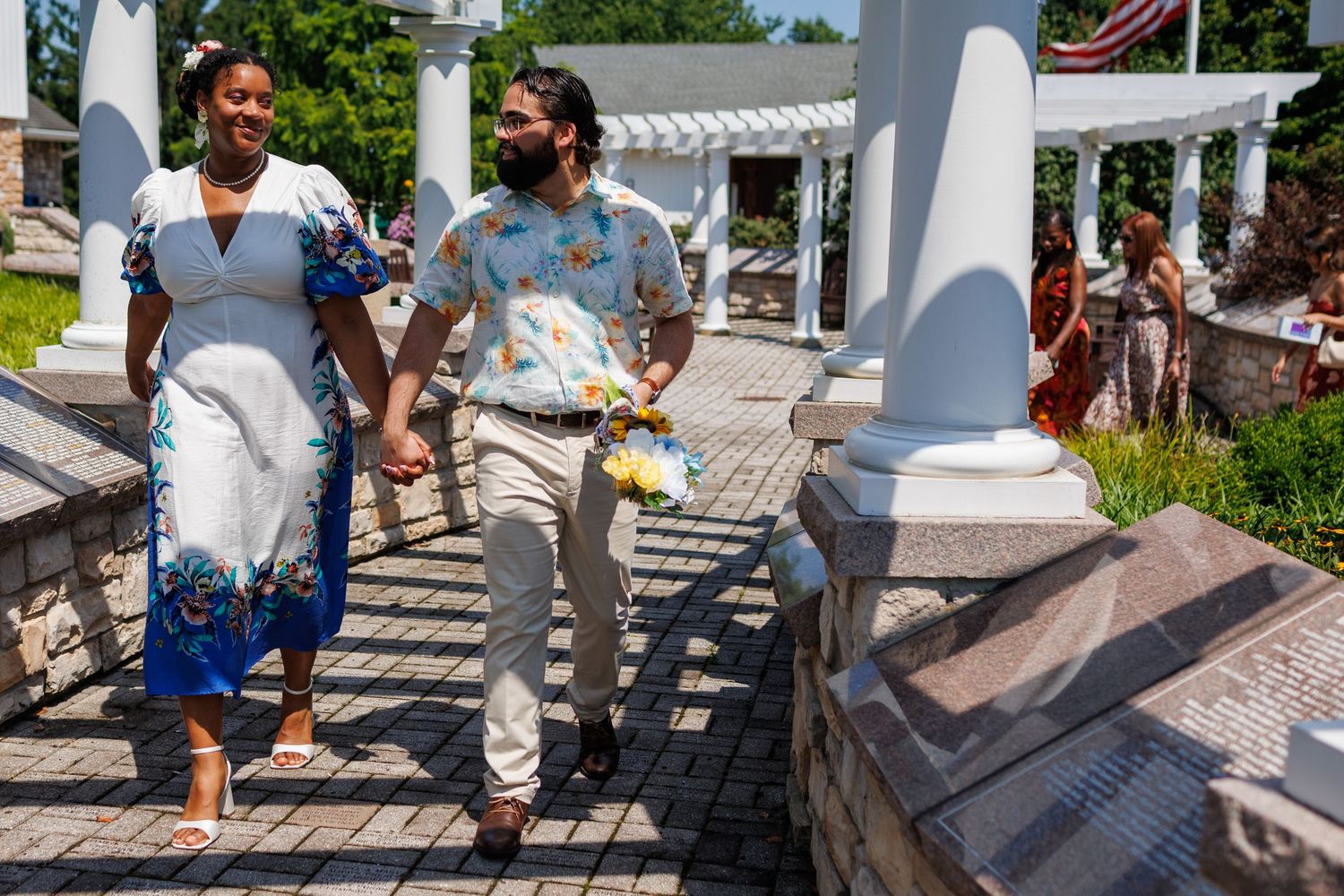 Couple in floral outfits and face masks walk hand-in-hand on a brick path at an outdoor wedding venue.