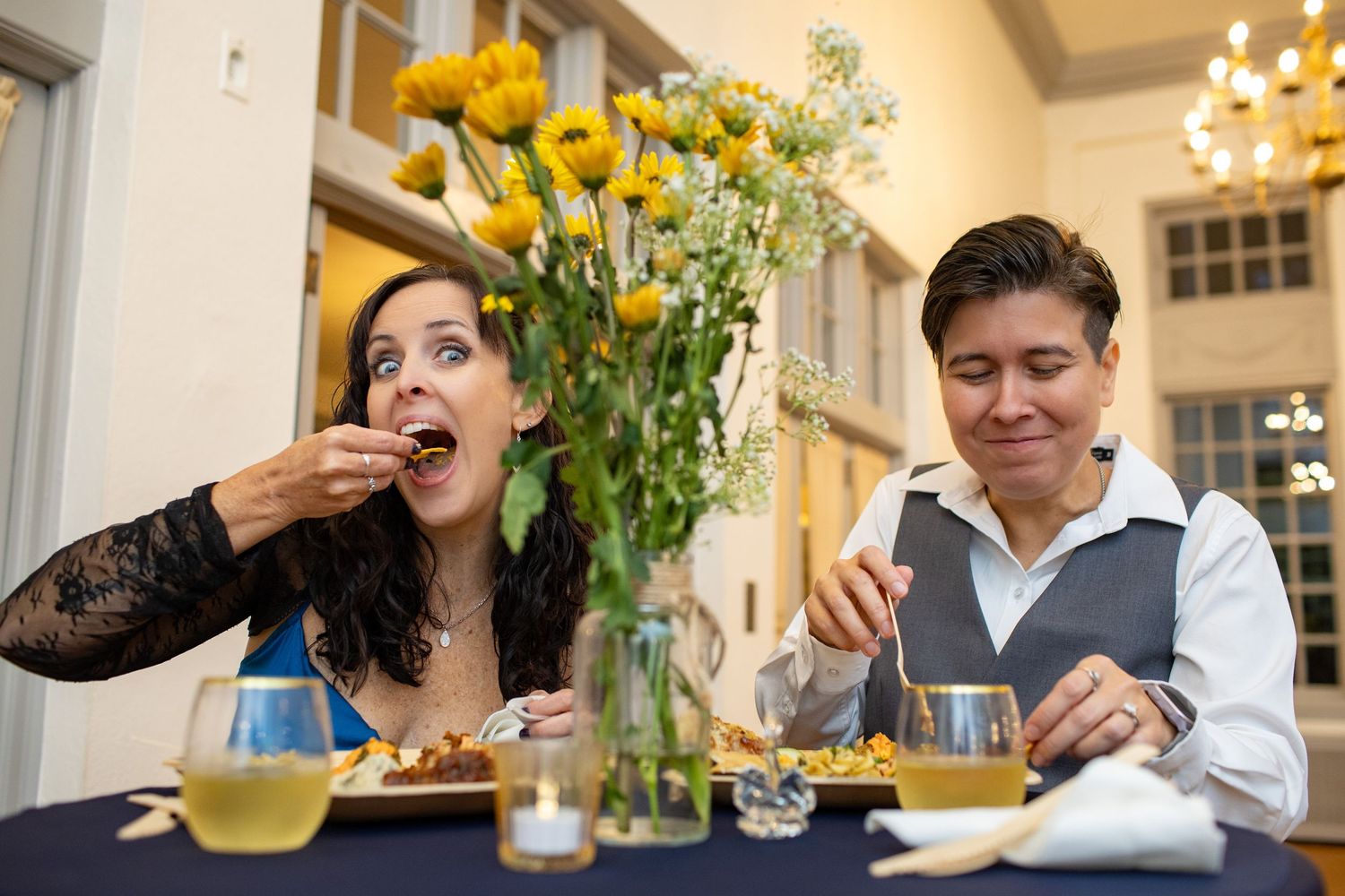 A group of people laughing and enjoying a meal together at a bright dining room table with yellow flowers.