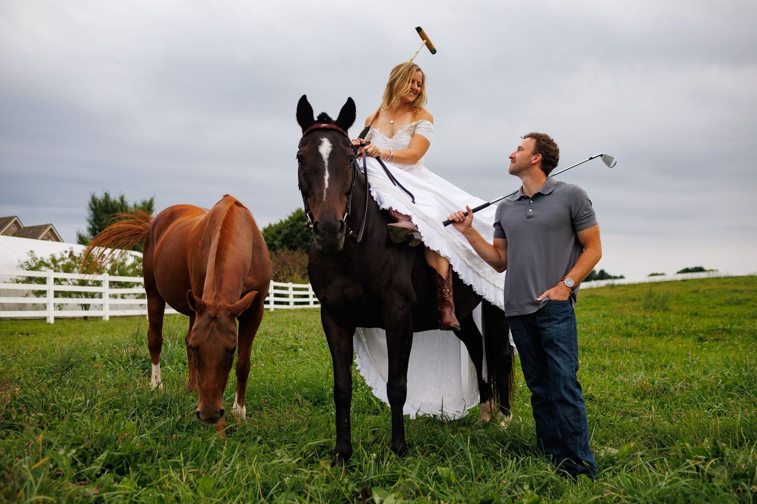Bride in white dress sitting on dark horse while groom holds golf club in green pasture with white fence.