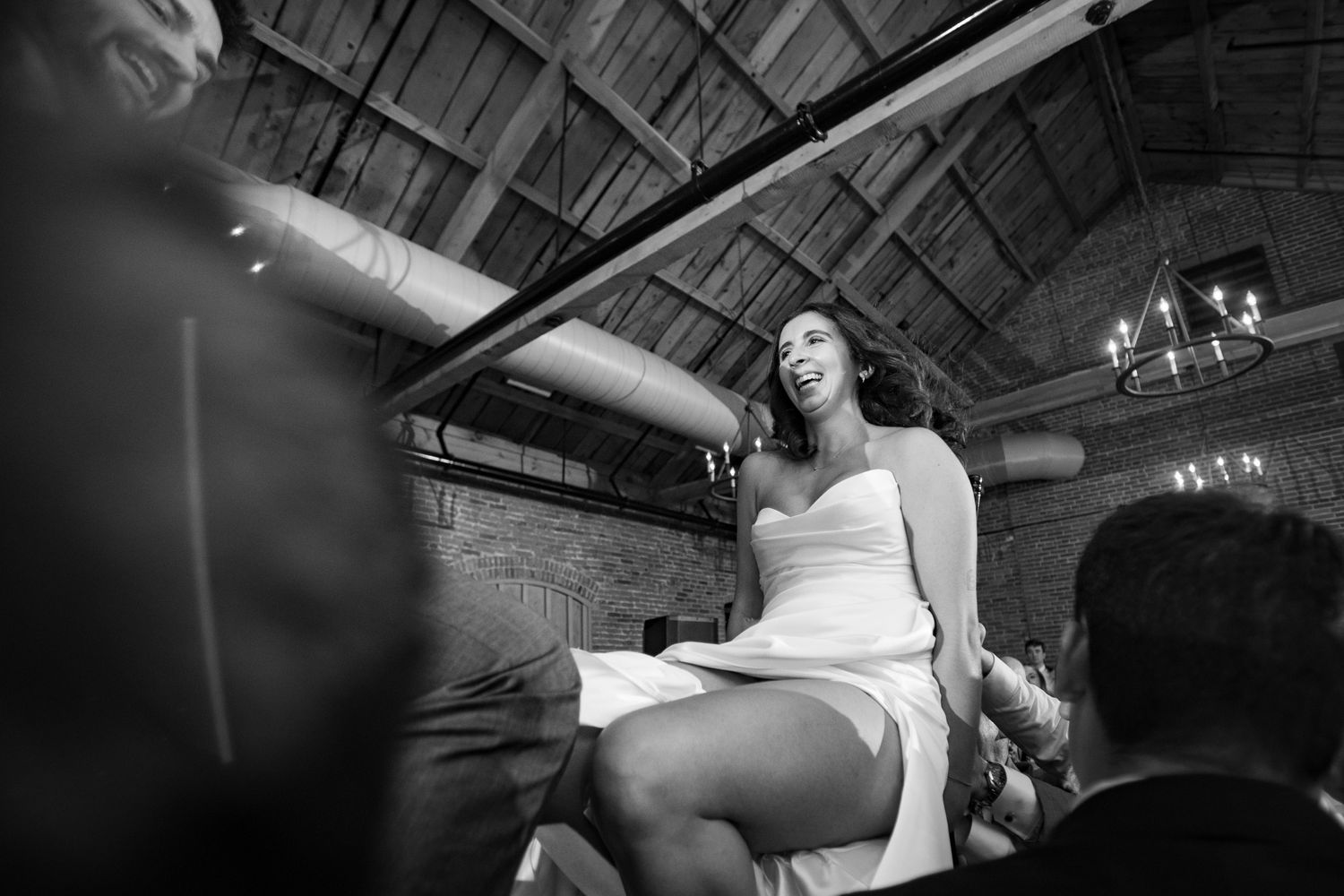 Black and white photo of woman in white dress being lifted by guests at wedding reception.
