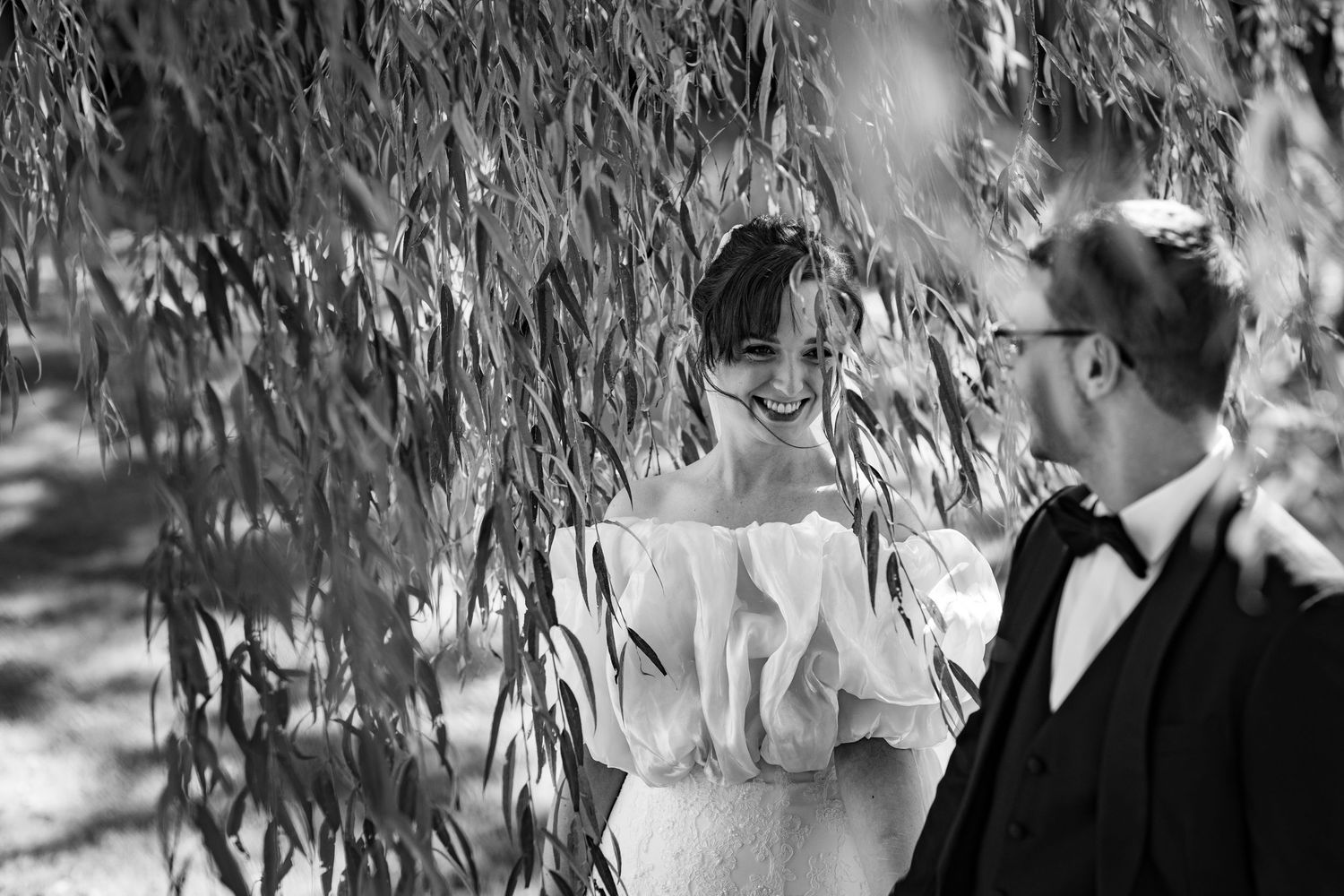 Bride and groom share a joyful moment under weeping willow tree in elegant black and white wedding photo.