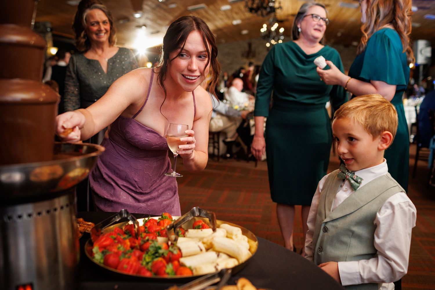 Woman and young boy in bow tie enjoying food at wedding reception chocolate fountain dessert table.