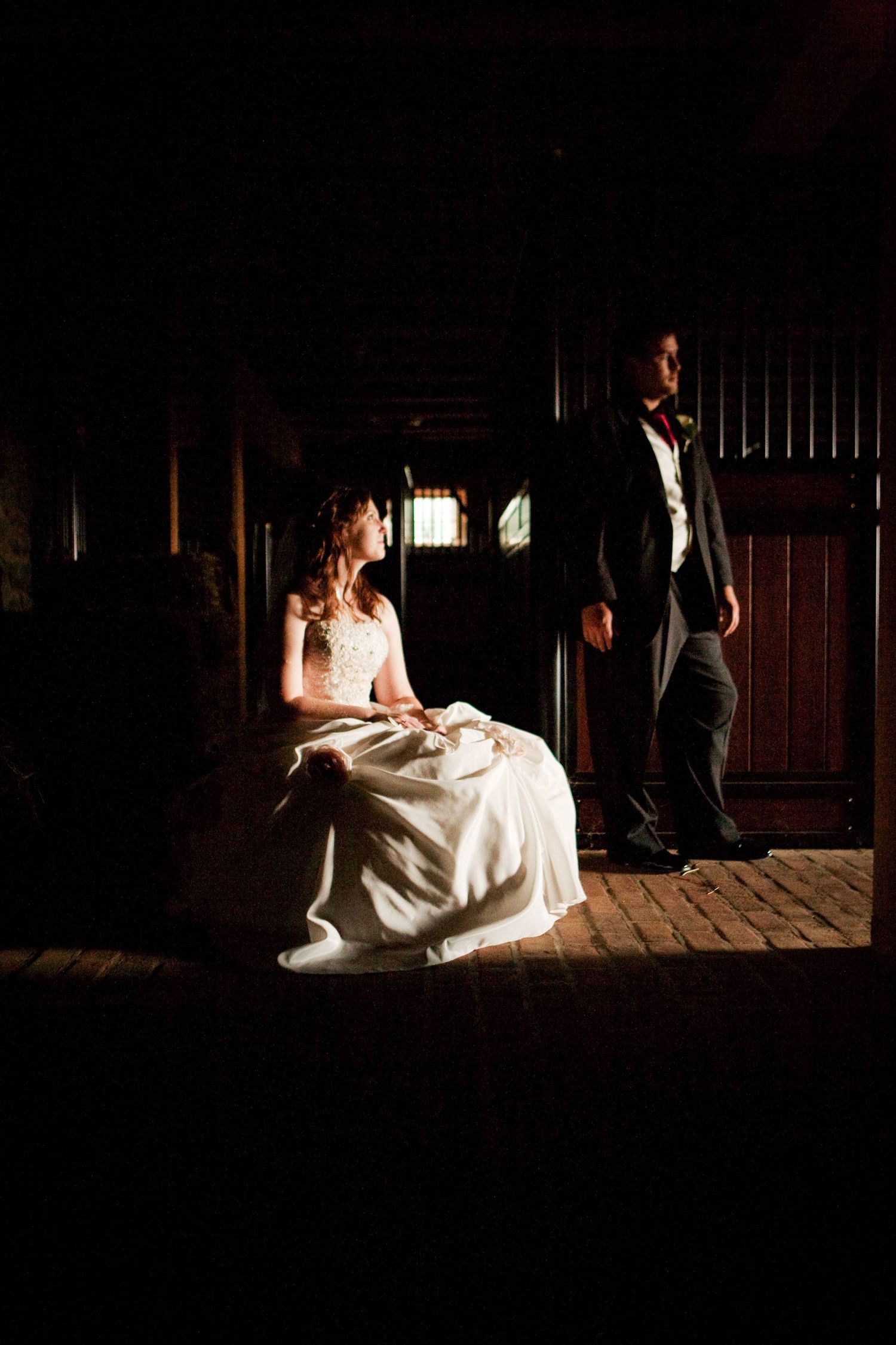 Dramatic lighting illuminates a bride seated in a rustic barn while a silhouetted figure stands in the shadows.