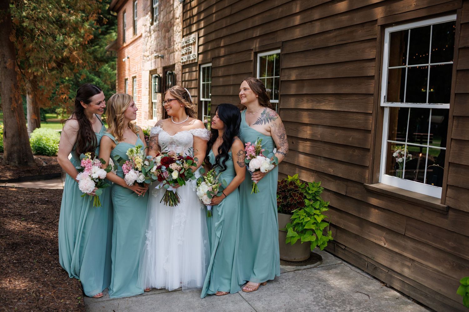 Bride in white gown smiles with four bridesmaids in sage green dresses holding bouquets outside rustic wooden building.