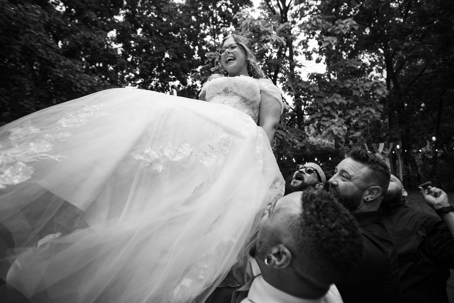 Black and white photo of joyful bride being lifted by wedding guests outdoors.