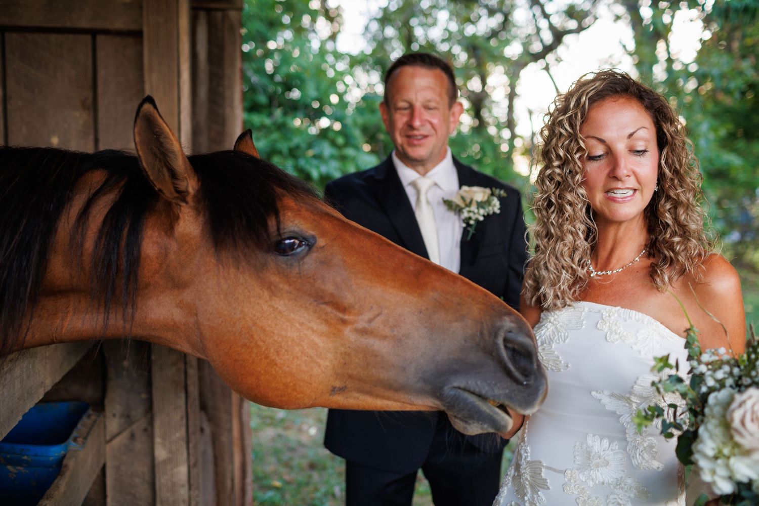 Bride in white strapless gown smiles as a brown horse nuzzles her, with groom in dark suit standing behind.