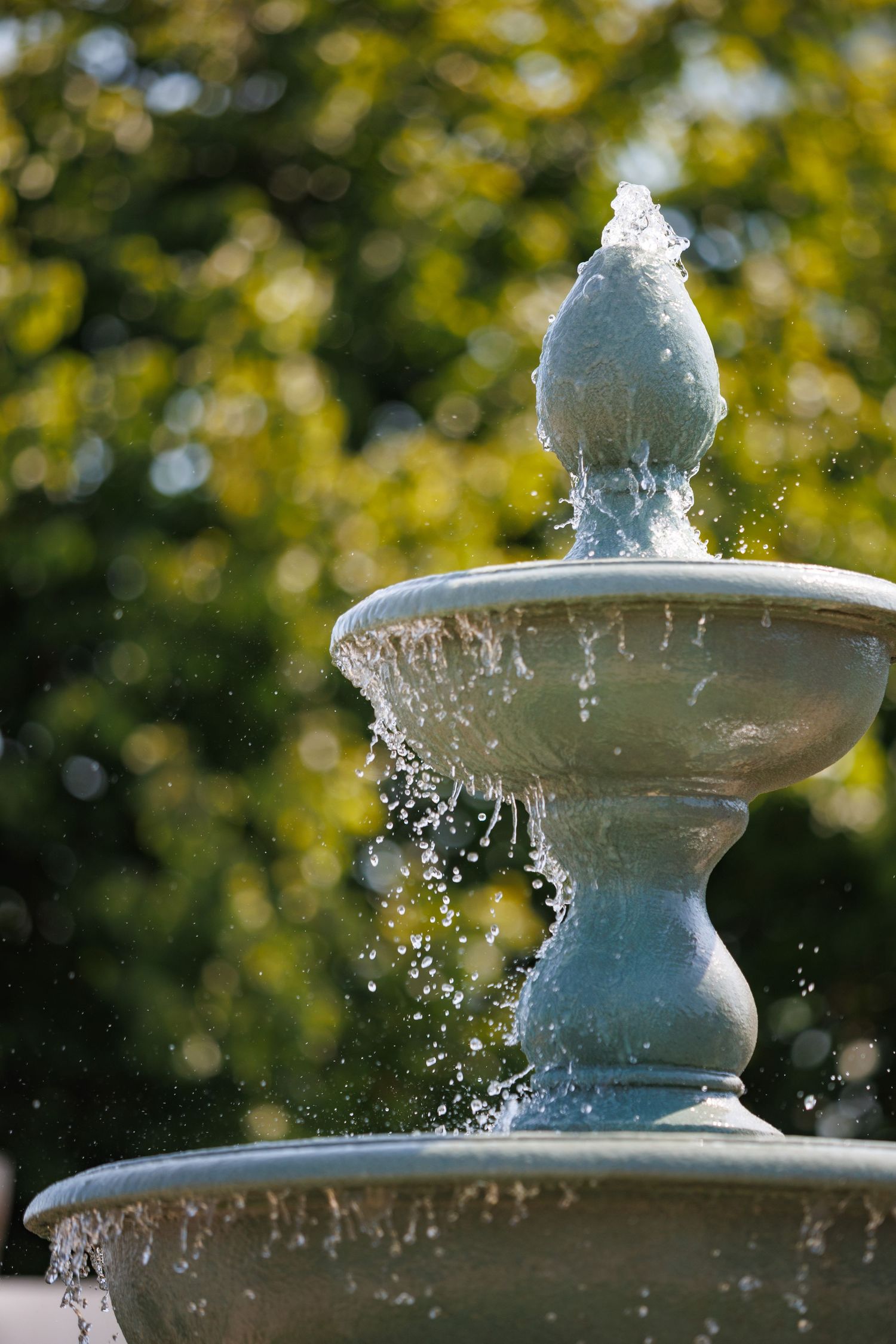A classic two-tiered stone garden fountain with water splashing, set against a lush green bokeh tree background.