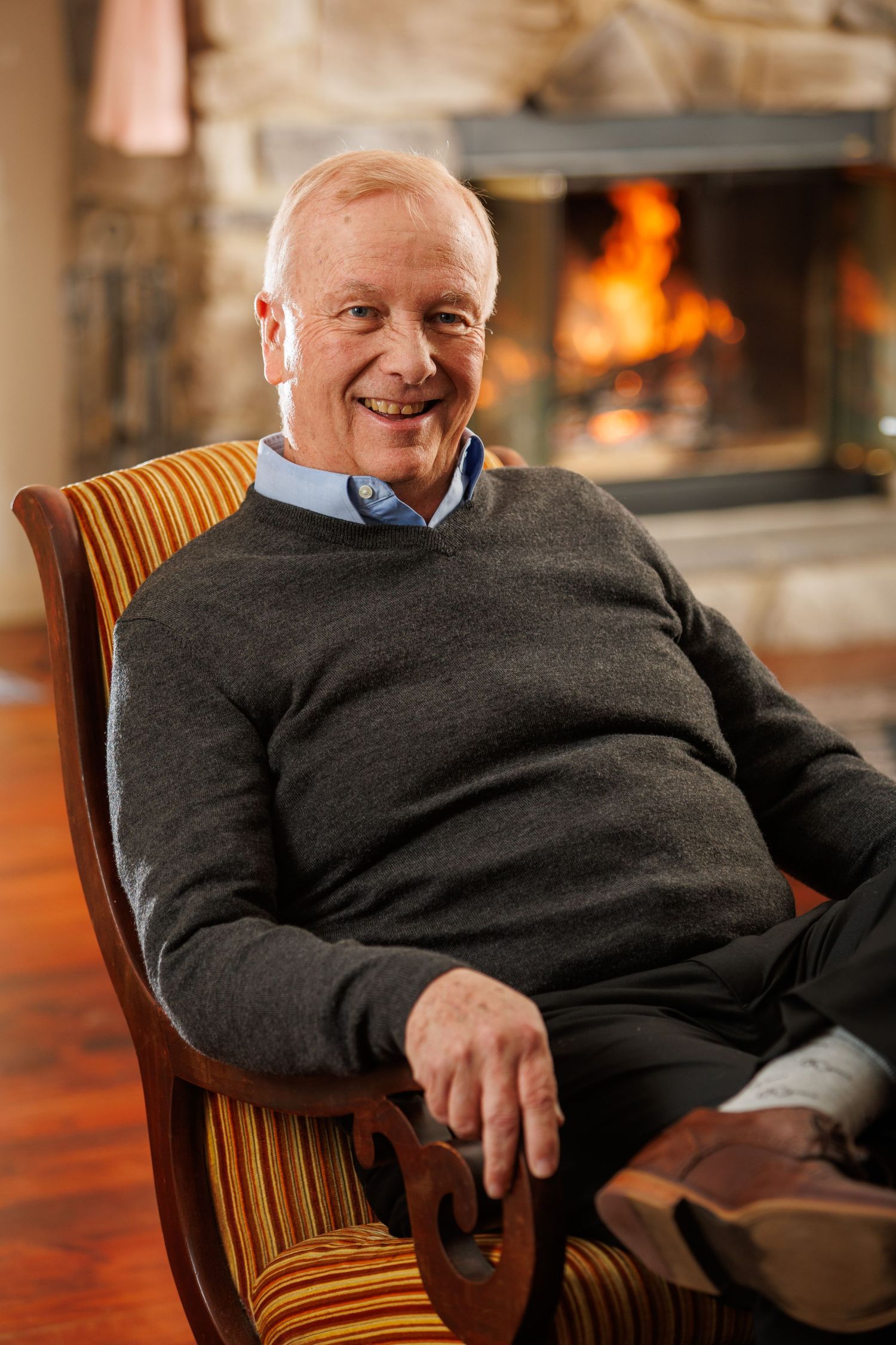 Person in dark sweater relaxes in wooden chair by warm fireplace with glowing flames.