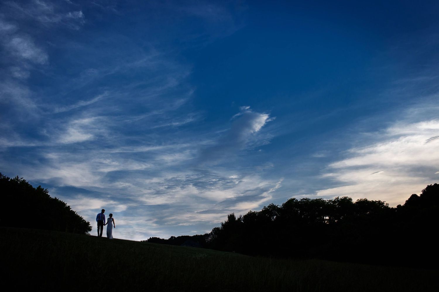 Silhouetted figures stand on hilltop against dramatic sunset sky.