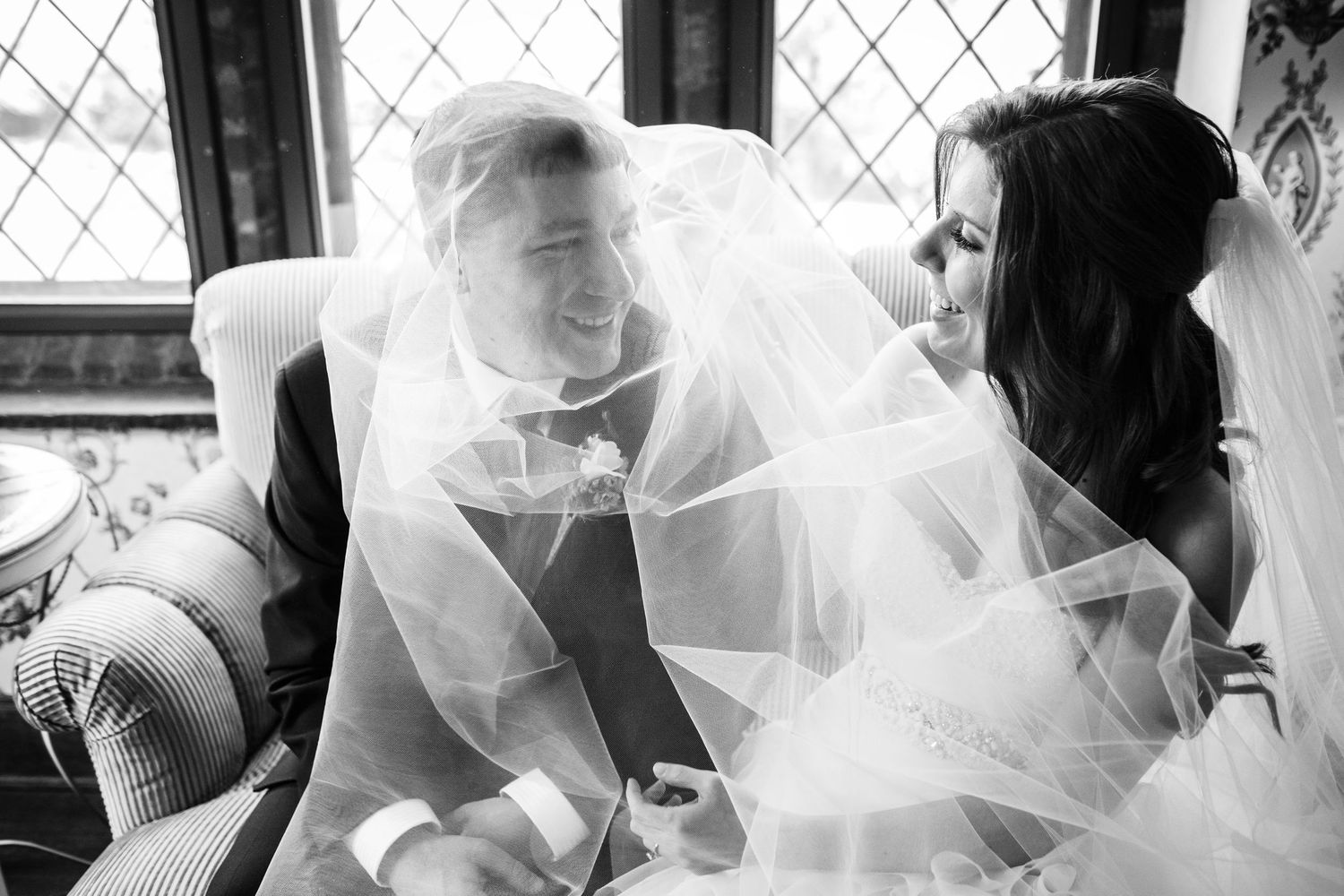 A joyful black and white wedding moment captured with the bride's veil flowing inside an ornate window setting.