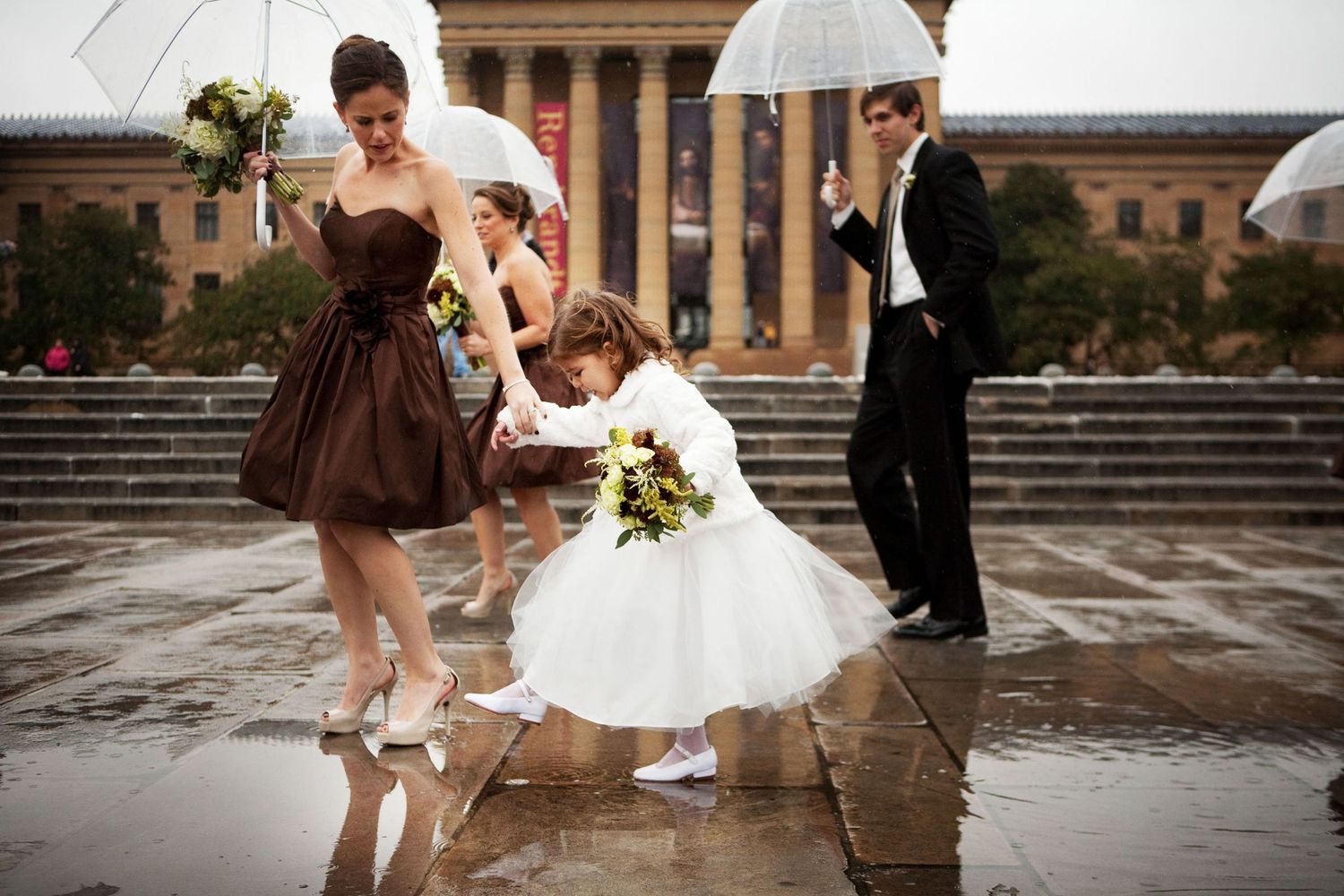 Bridal party playfully splashing in puddles on rainy day with umbrellas outside historic building steps.