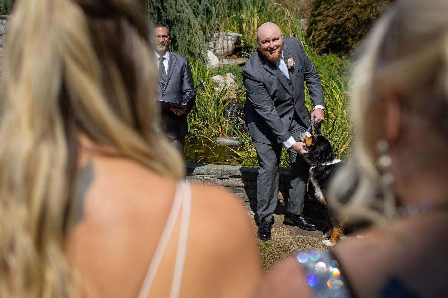 A wedding couple stands in a sunny garden surrounded by greenery while guests look on in the background.