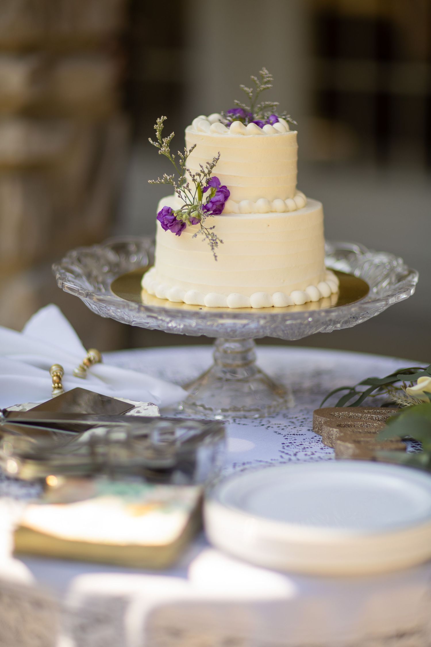 Close-up views of a round white wedding cake with purple flowers displayed on a delicate lace-edged cake stand.