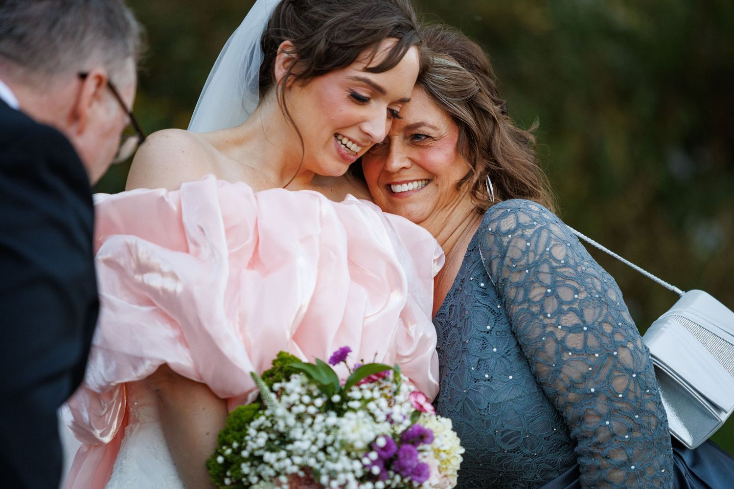 Emotional bride in pink gown shares tender hug with older woman holding baby's breath bouquet.