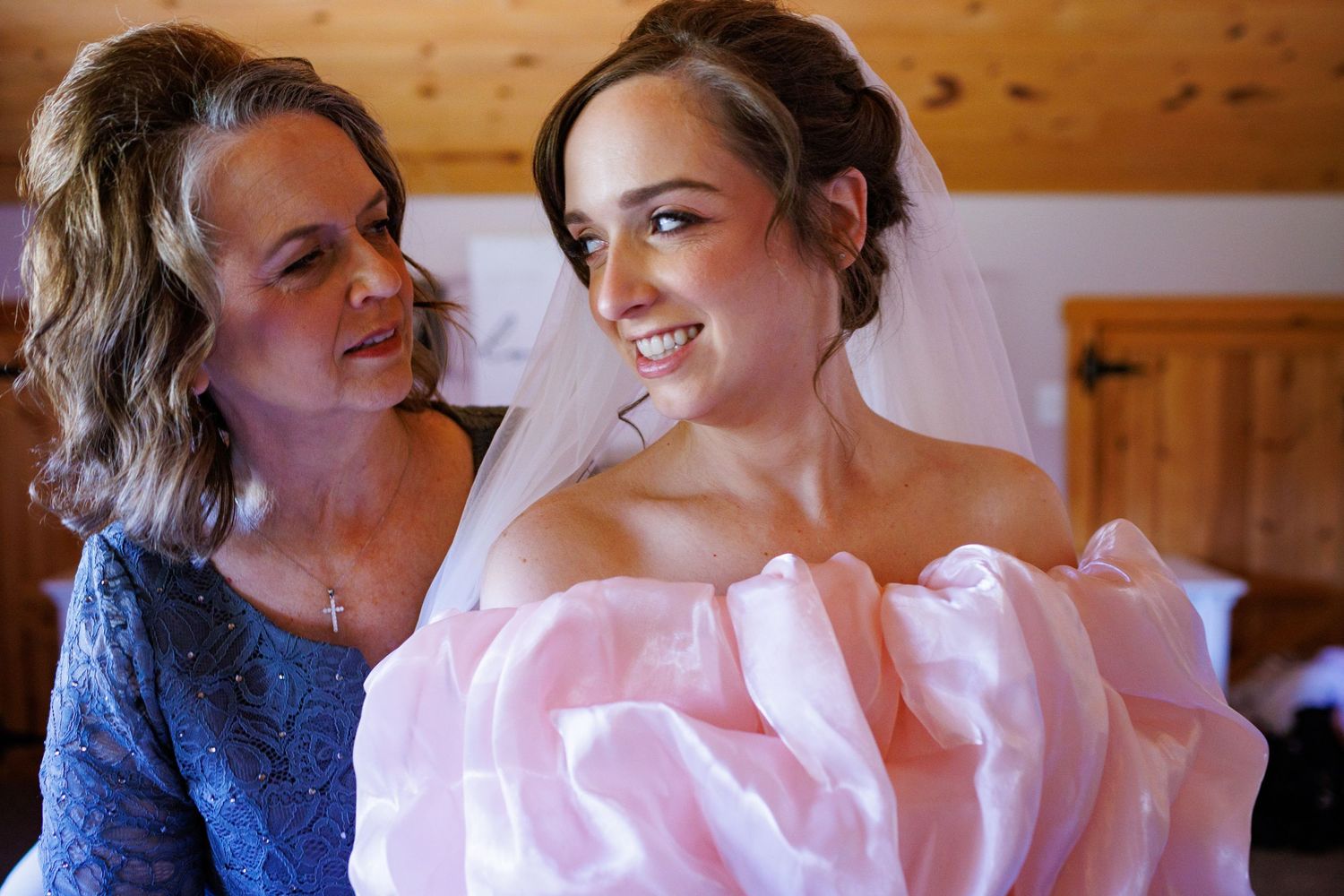 Mother helping bride into a pink wedding dress, both smiling joyfully before the ceremony.