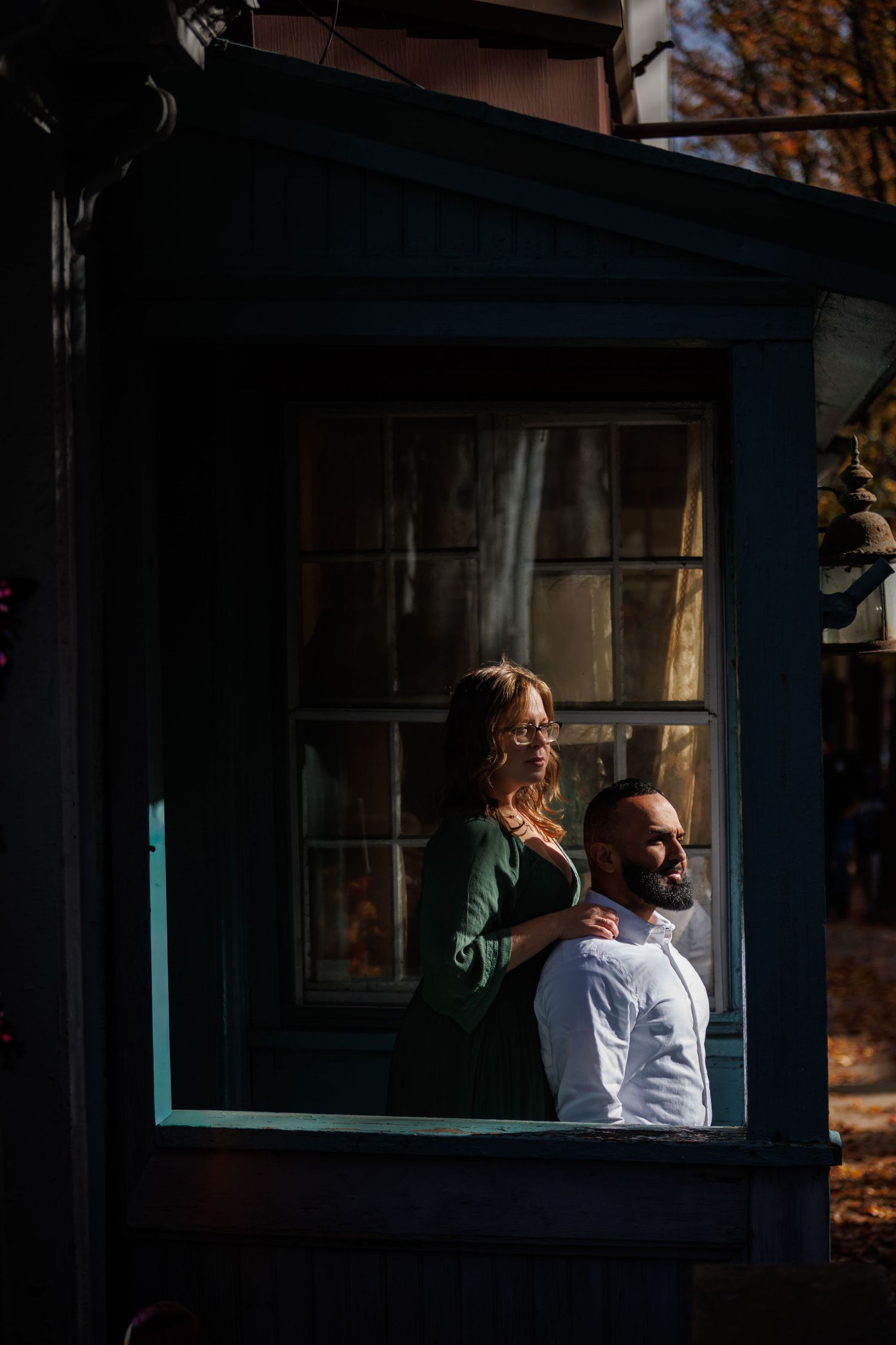 Two engaged people sit together in a dark window frame with sunlight casting dramatic shadows and highlights.