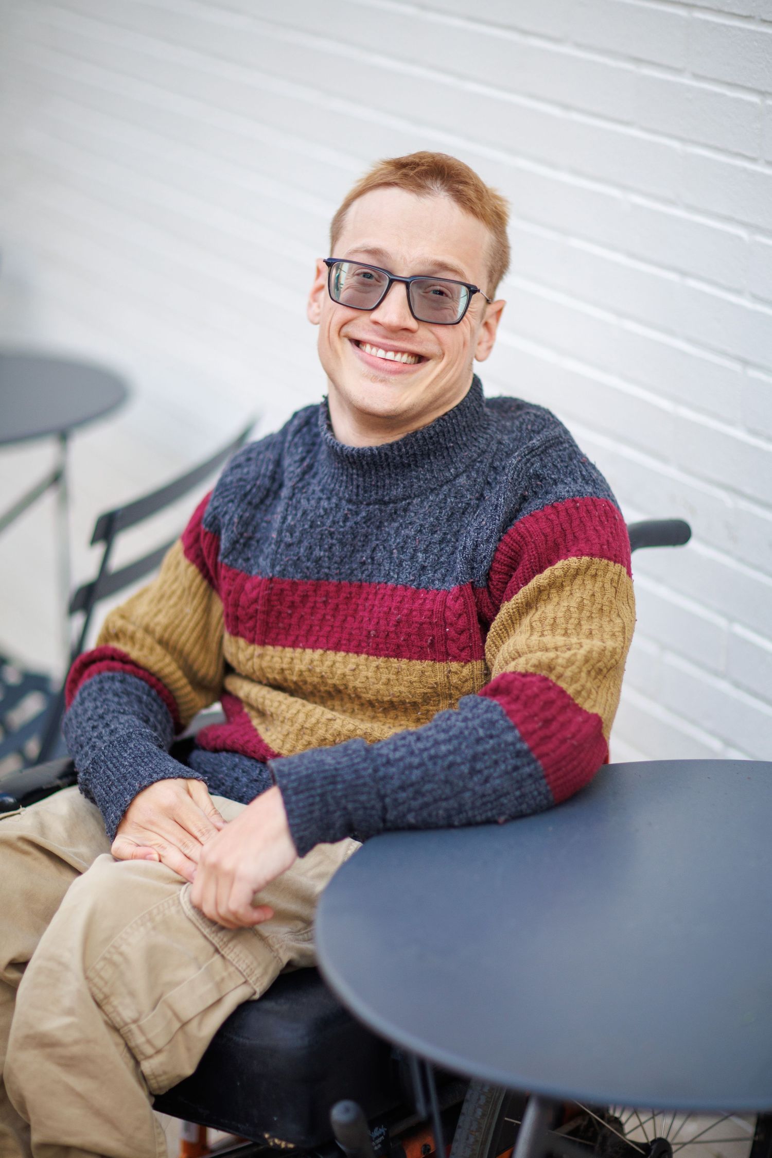 Someone wearing glasses and a colorful striped sweater sits at a round table against a white brick wall.