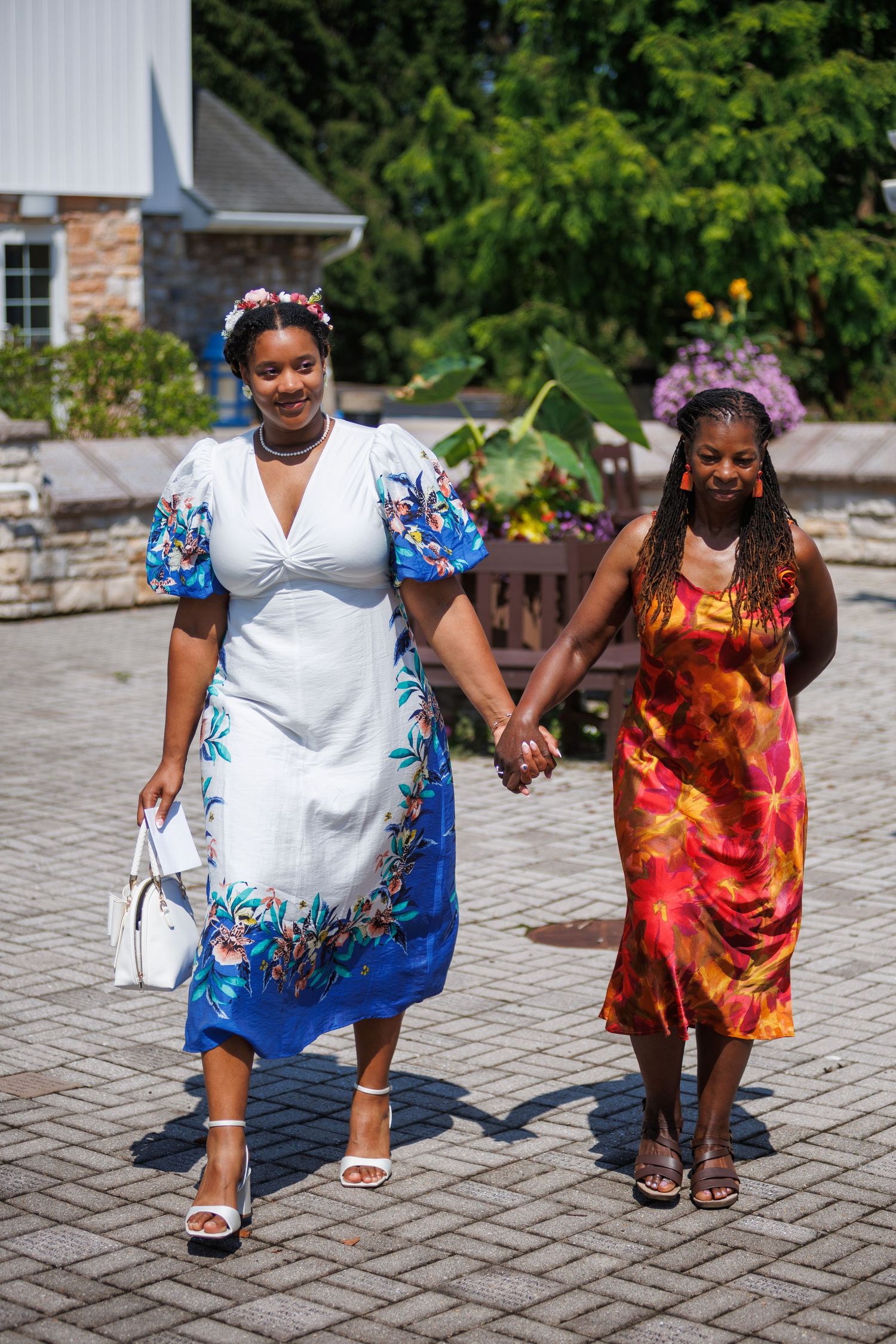 Two women holding hands walking outdoors, one in a floral white dress with flower crown, other in tie-dye dress.