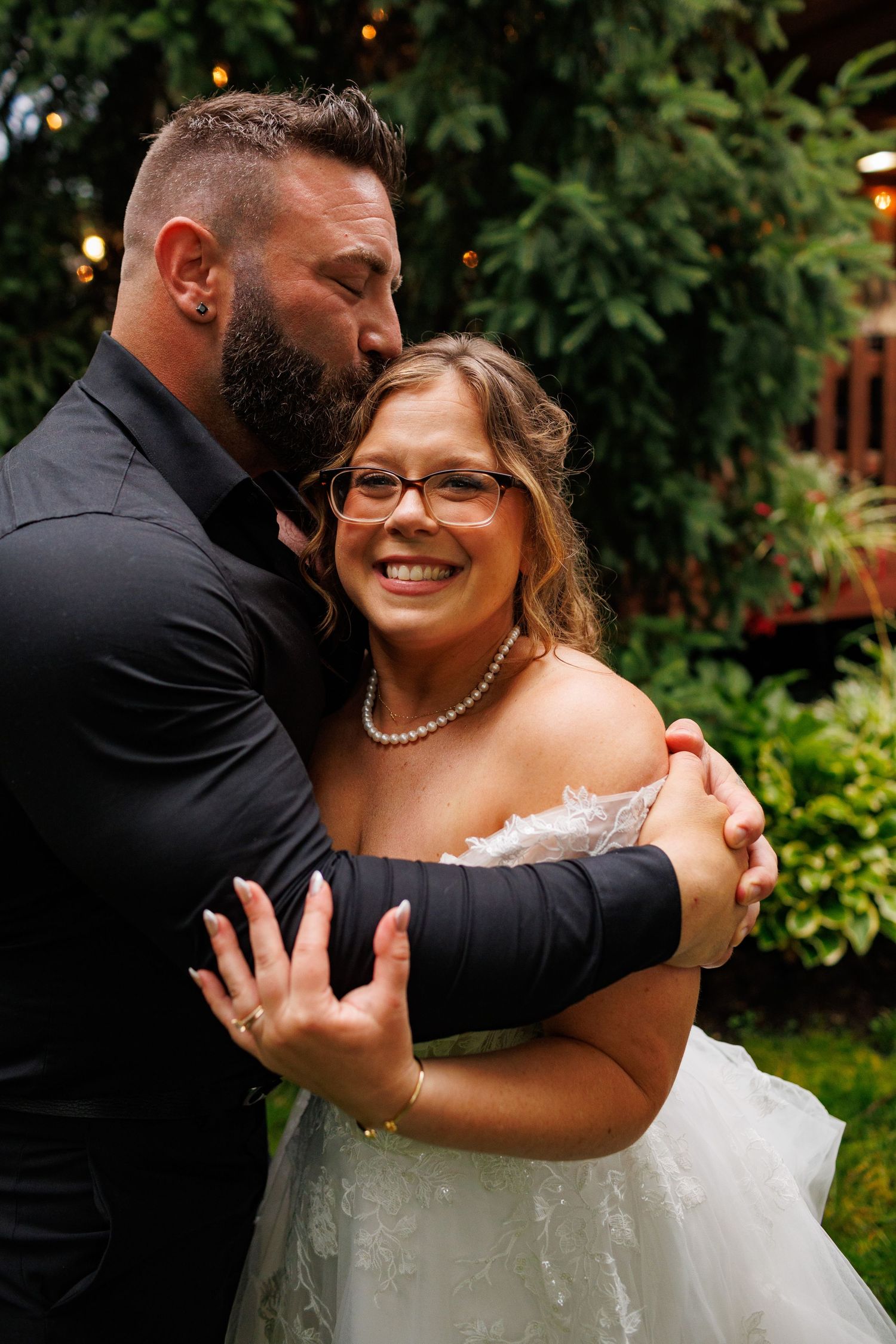 Bearded groom in black shirt embracing smiling bride in white gown outdoors at wedding.