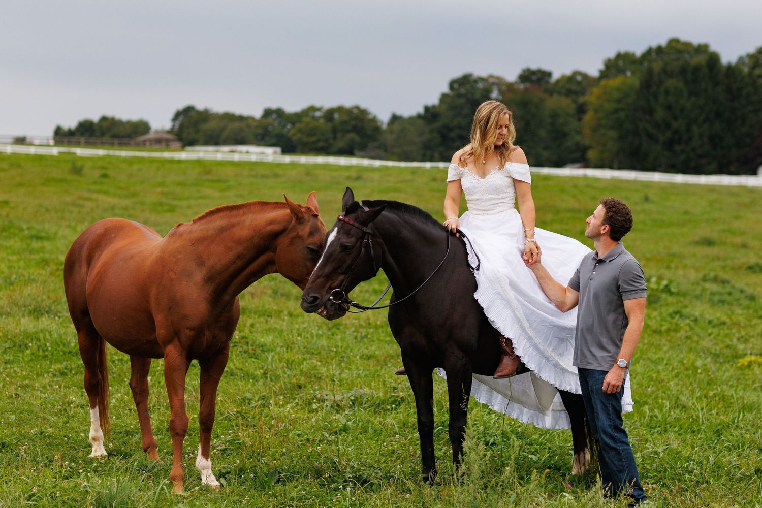 Bride in white gown sits atop a dark horse beside a chestnut horse as groom holds her dress in a green field.
