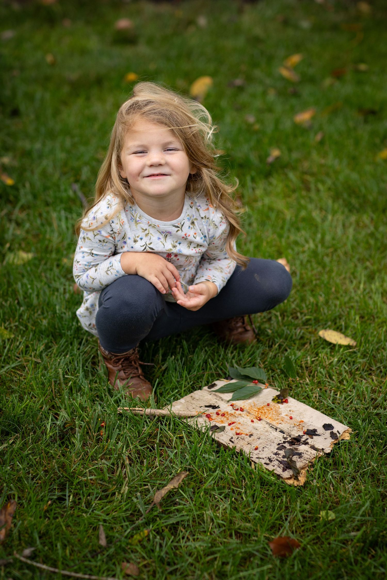 A child in a floral shirt crouches on grass near a weathered piece of wood on an autumn day.