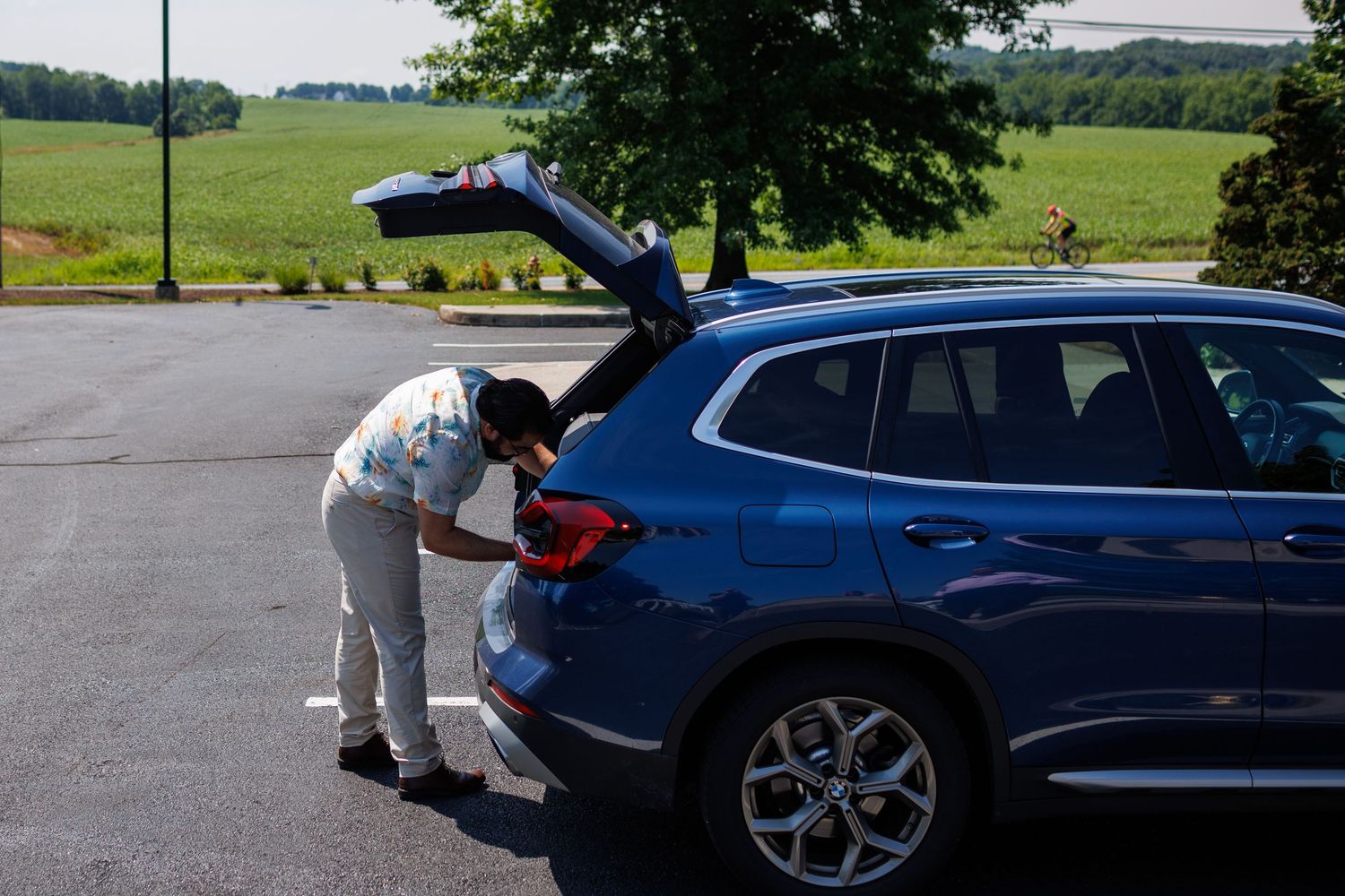Person loading items into the trunk of a blue BMW SUV in a parking lot surrounded by green trees.