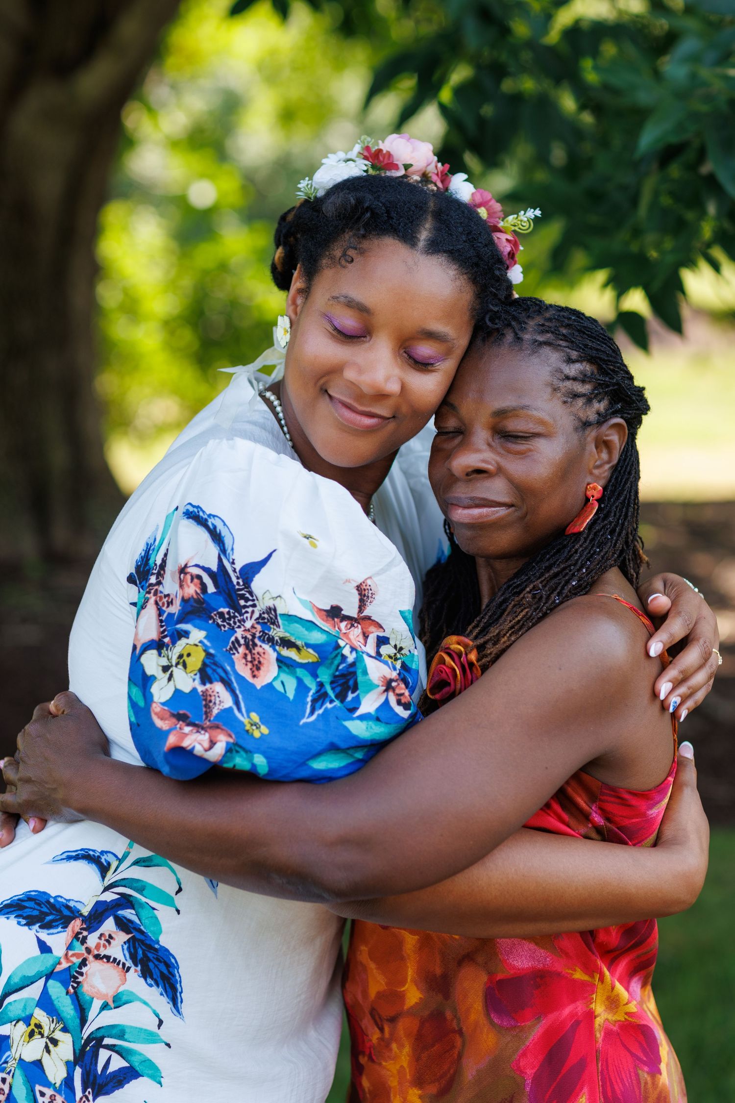Two women sharing a warm embrace outdoors, one wearing a floral dress and flower crown, smiling joyfully.