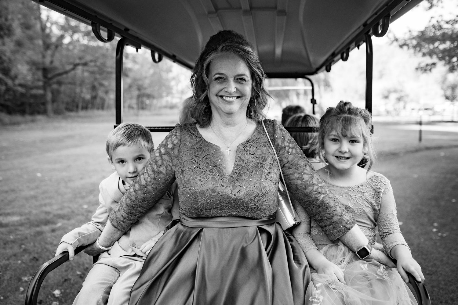 A smiling woman in a lace dress rides a golf cart with two young children at a wedding.
