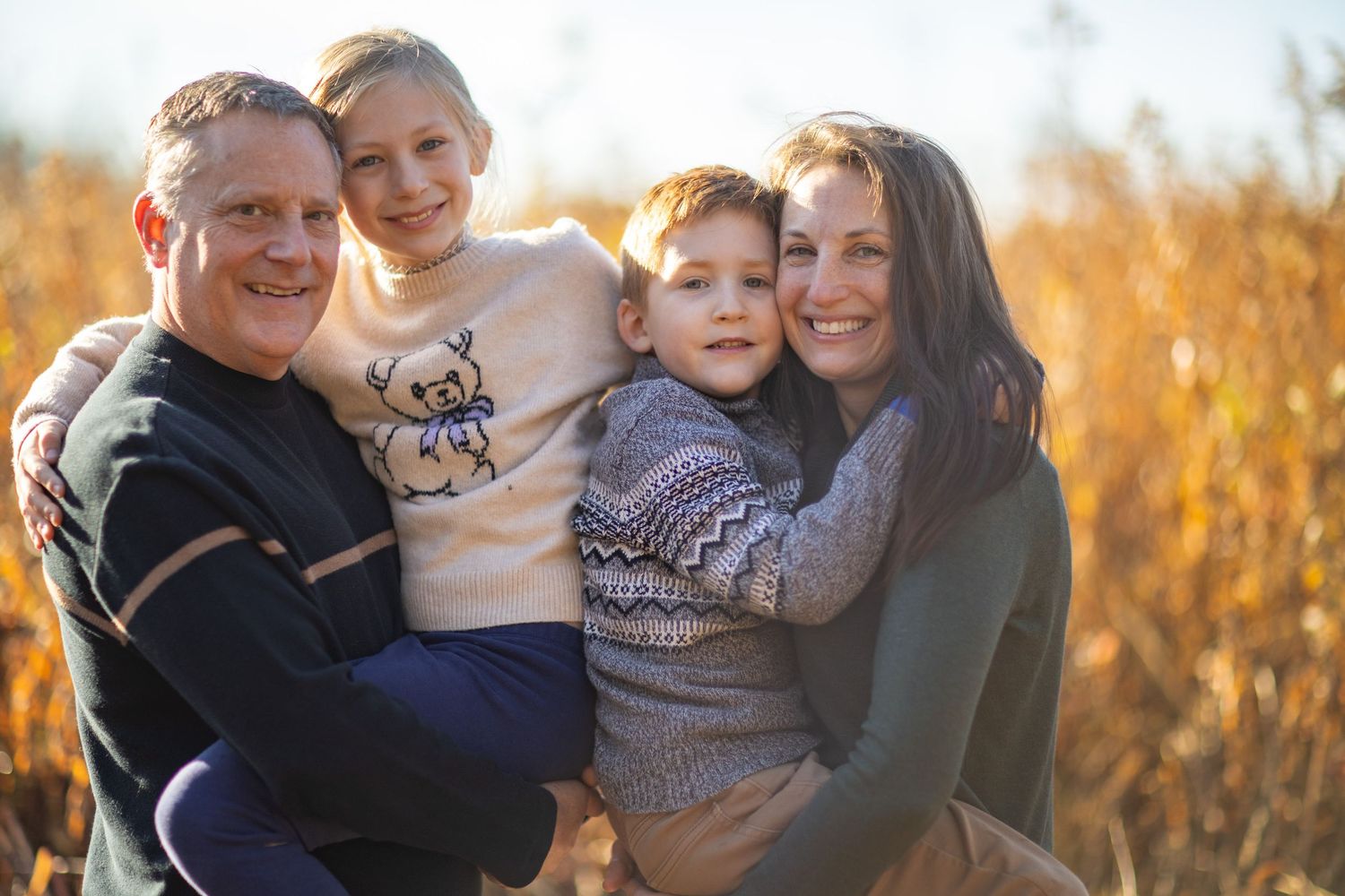 A warm family portrait session outdoors with golden sunlight filtering through autumn trees in the background.