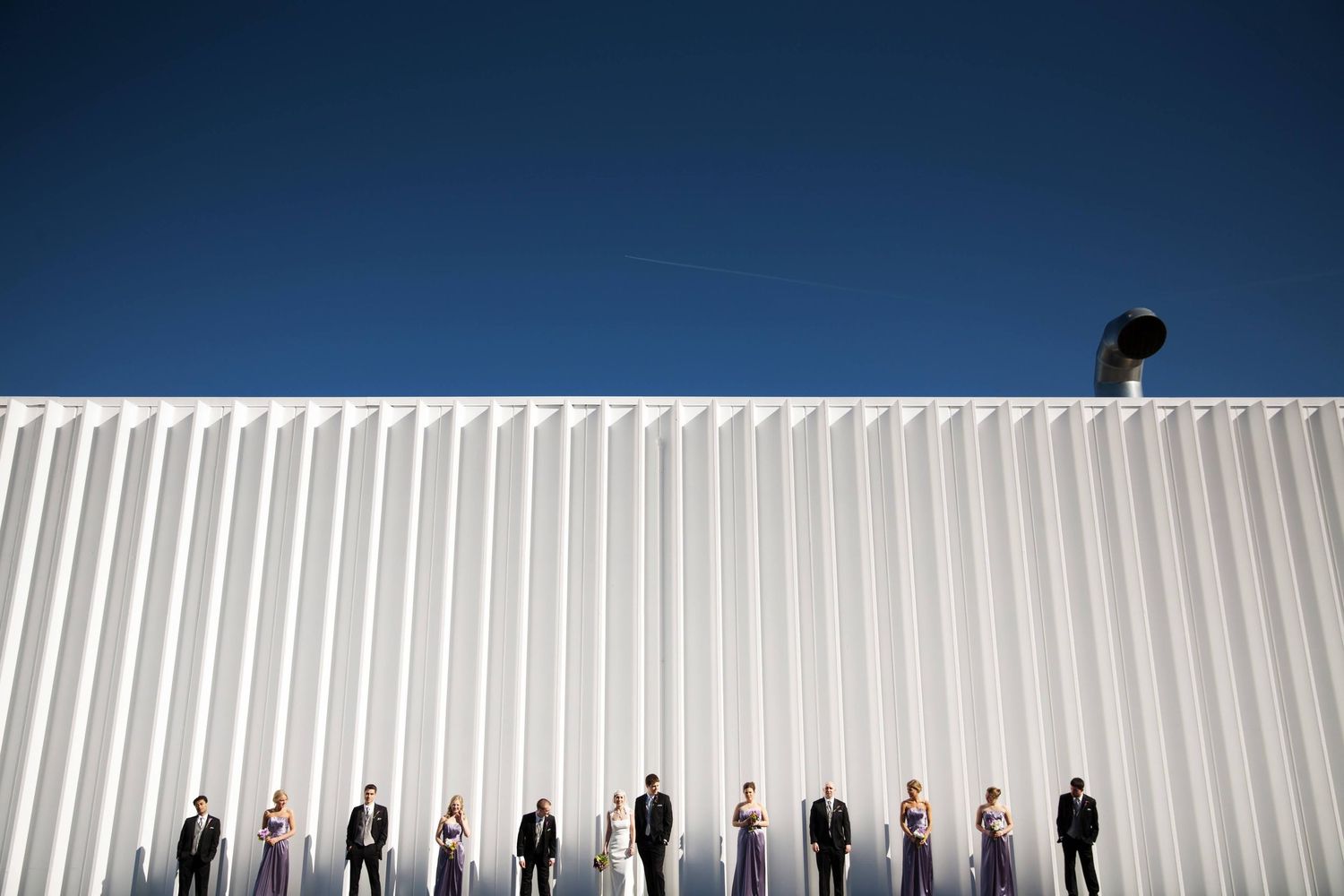 Wedding party lined up against white industrial wall under blue sky.