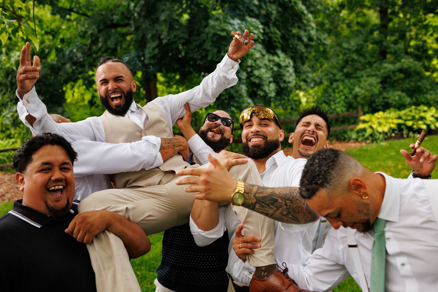 Groom carried by laughing groomsmen in white attire celebrating outdoors after wedding ceremony.