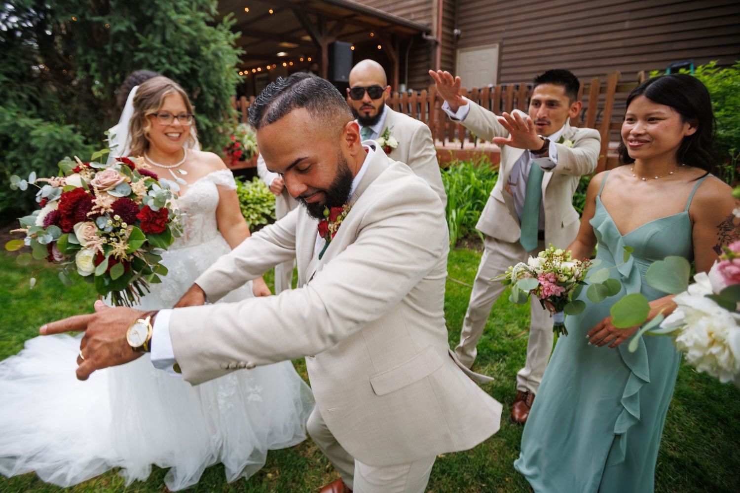 Groom in tan suit celebrates joyfully with wedding party in garden, bridesmaids in sage green holding floral bouquets.
